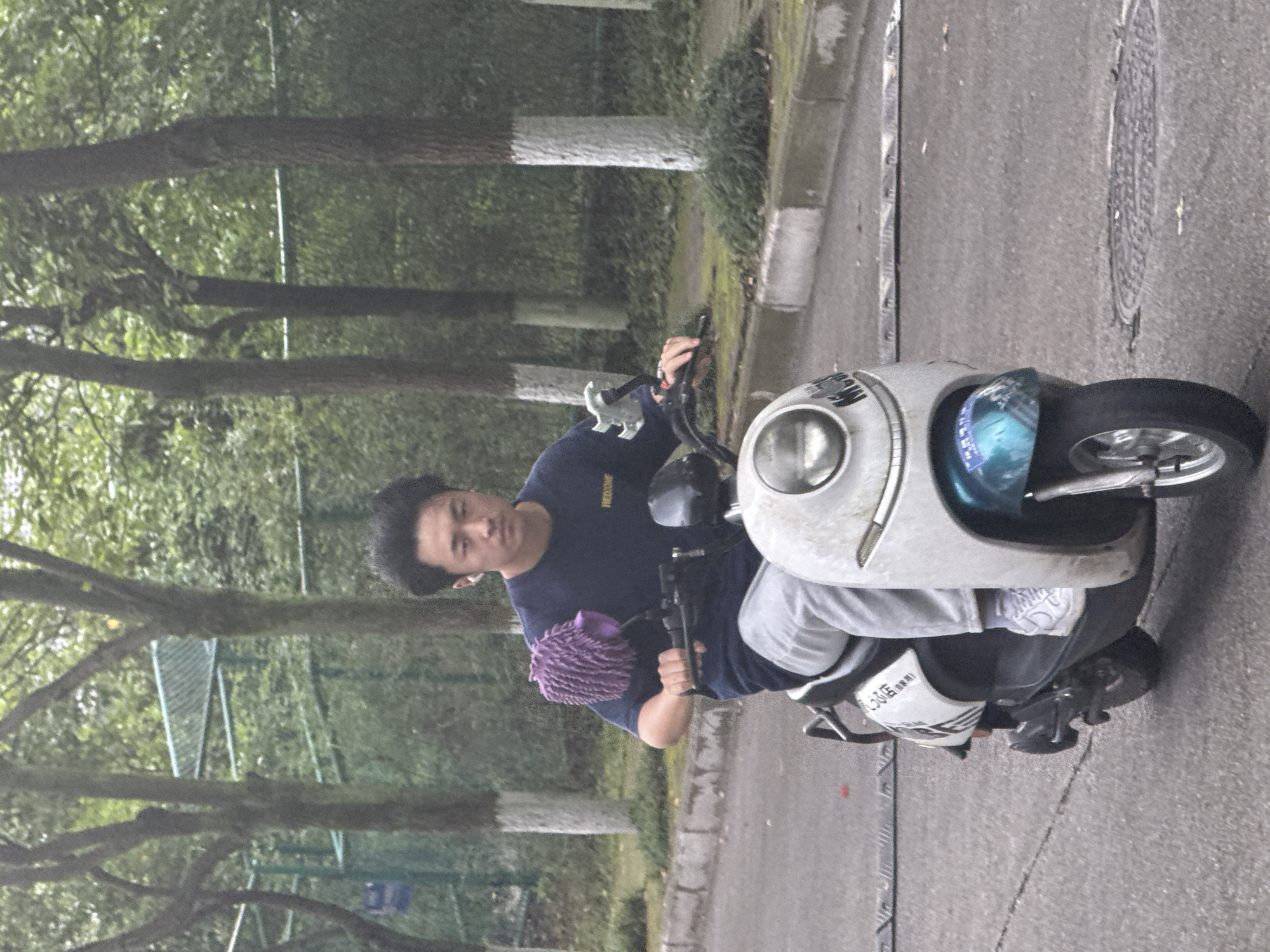 A medium shot shows a person riding a white scooter on a paved road, the location appears to be an urban area in the city of Jiangjin, China. The scooter has a round headlight and a blue-tinted windshield. The rider wears a dark blue t-shirt, light-colored pants, and black shoes. A purple cleaning mitt is attached to the scooter's handlebar. The background includes trees, a metal fence, and other greenery.