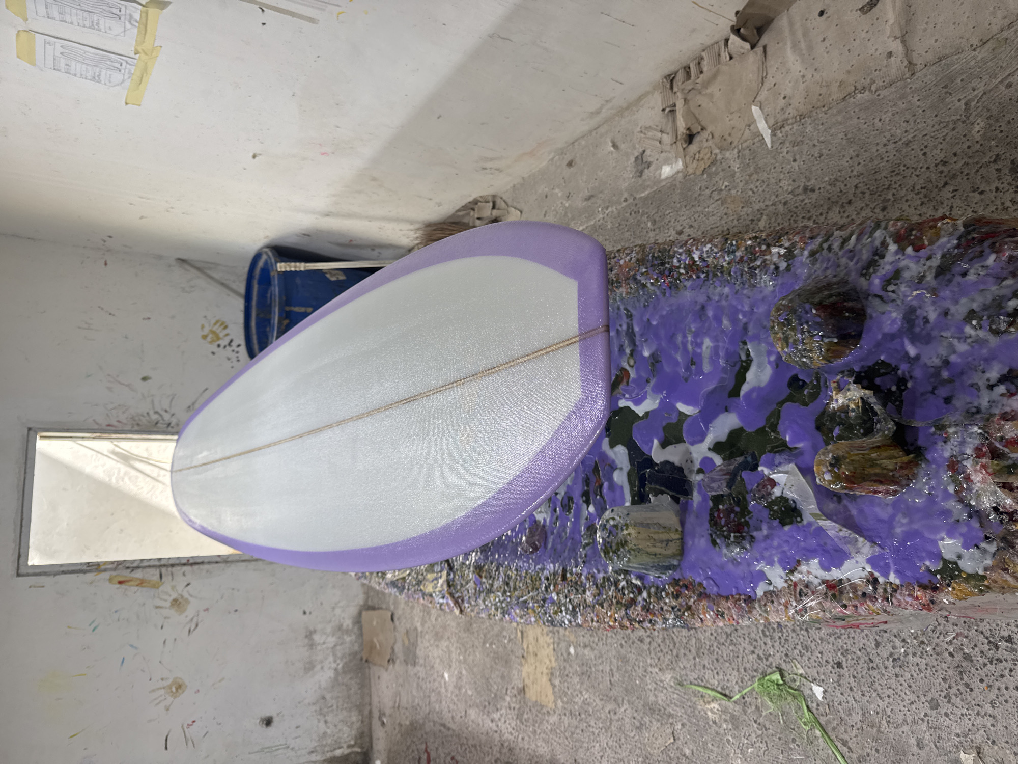 The image depicts a surfboard resting on a cluttered work surface. The surfboard has a white body with a purple trim. The work surface is covered in a mix of colorful materials, with a predominance of purple and white. The surroundings appear to be an indoor workspace with textured walls. A blue barrel is visible in the background, and there are hints of a window or doorway in the lower-left corner. Given the prompt mentions Denpasar, Indonesia, and the presence of what appears to be surfboard construction materials, the image may depict a surfboard workshop in that city.