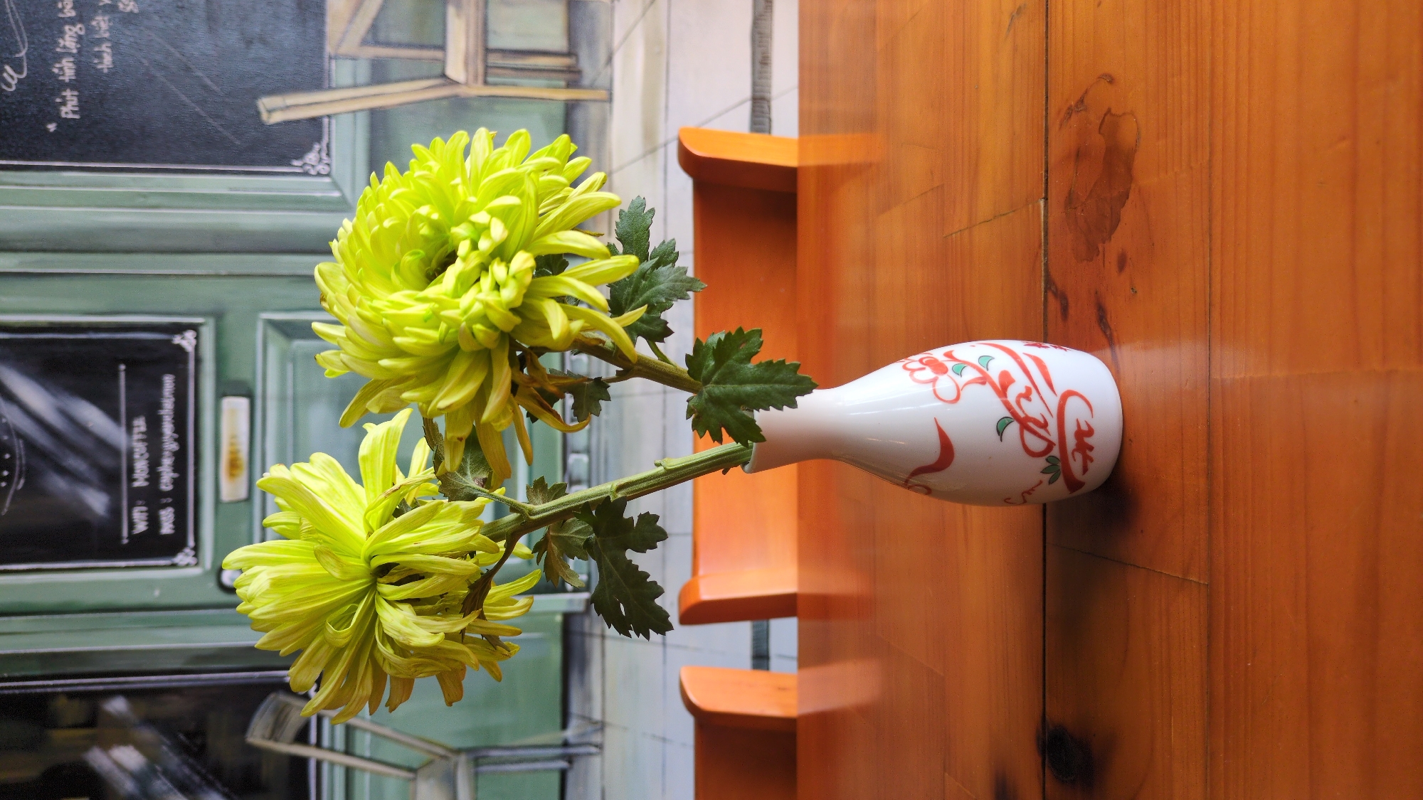 On a wooden table indoors, two yellow chrysanthemum flowers with green leaves sit in a white vase with red floral designs. The vase is horizontal, on its side. Behind the flowers and vase, a tiled wall with an abstract painting of a building is visible. The scene is photographed in the city of Da Lat, Vietnam.