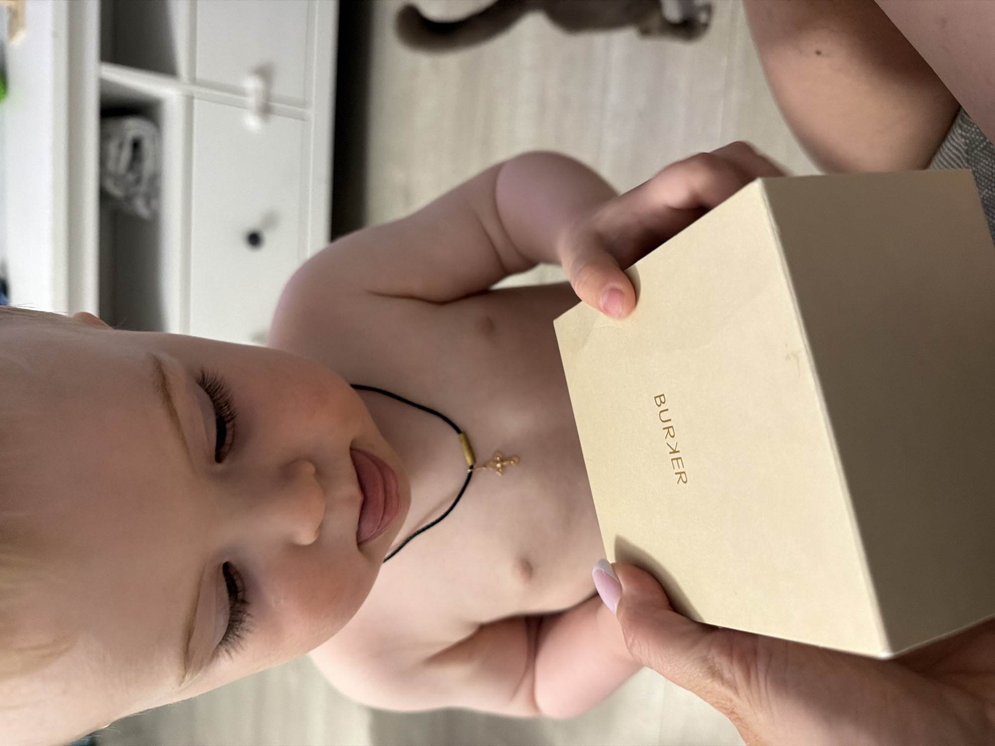 A baby with fair skin is partially visible, looking down and sticking its tongue out, wearing a black cord necklace with a gold cross. The baby is positioned to the left of a rectangular, light-colored box with "BURKER" printed vertically in gold lettering on the side. A hand with a light pink nail polish is holding the corner of the box. A piece of furniture, possibly a shelf, is visible on the left, and a cat's tail is visible in the background. The location is unknown.