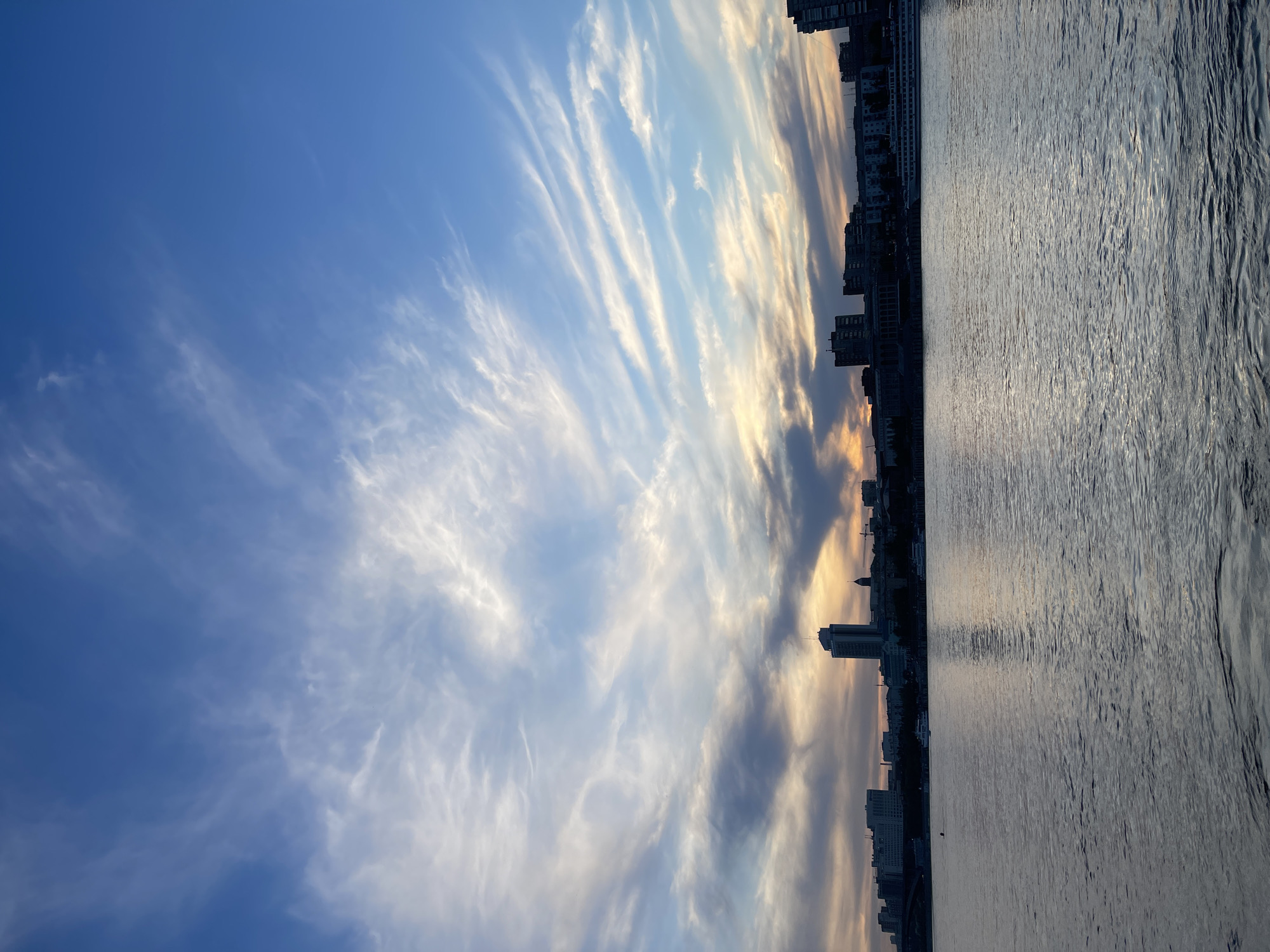 The image displays a serene panoramic view of a cityscape along a wide body of water, likely a river, under a dramatic sky. The horizon line runs horizontally across the middle of the frame.
The sky occupies the upper two-thirds of the image, showcasing a transition of colors. On the left, it is a deep, clear blue, gradually lightening and blending into soft blues, whites, and warm golden-orange tones towards the right side. This color palette strongly suggests either sunrise or sunset. Wispy, elongated clouds, likely cirrus or cirrocumulus, stretch across the sky, particularly concentrated in the middle and right portions, illuminated by the low-lying sun. These clouds create striking patterns, with some appearing bright white and others tinged with gold or soft grey.
Along the horizon, a silhouette of urban buildings forms the city skyline. These are predominantly tall structures, appearing dark against the lighter sky, indicating a significant urban presence. Below the city and sky, the wide expanse of water fills the bottom third of the image. The water's surface is relatively calm, with gentle ripples visible, especially towards the right. It reflects the muted light of the sky and the dark shapes of the city, with subtle glimmers of reflected light scattered across its surface.
The scene captures a tranquil moment during the "golden hour" in a major city like Krasnoyarsk, Russia, emphasizing the vastness of the sky and water against the urban development. There is no visible text in the image.