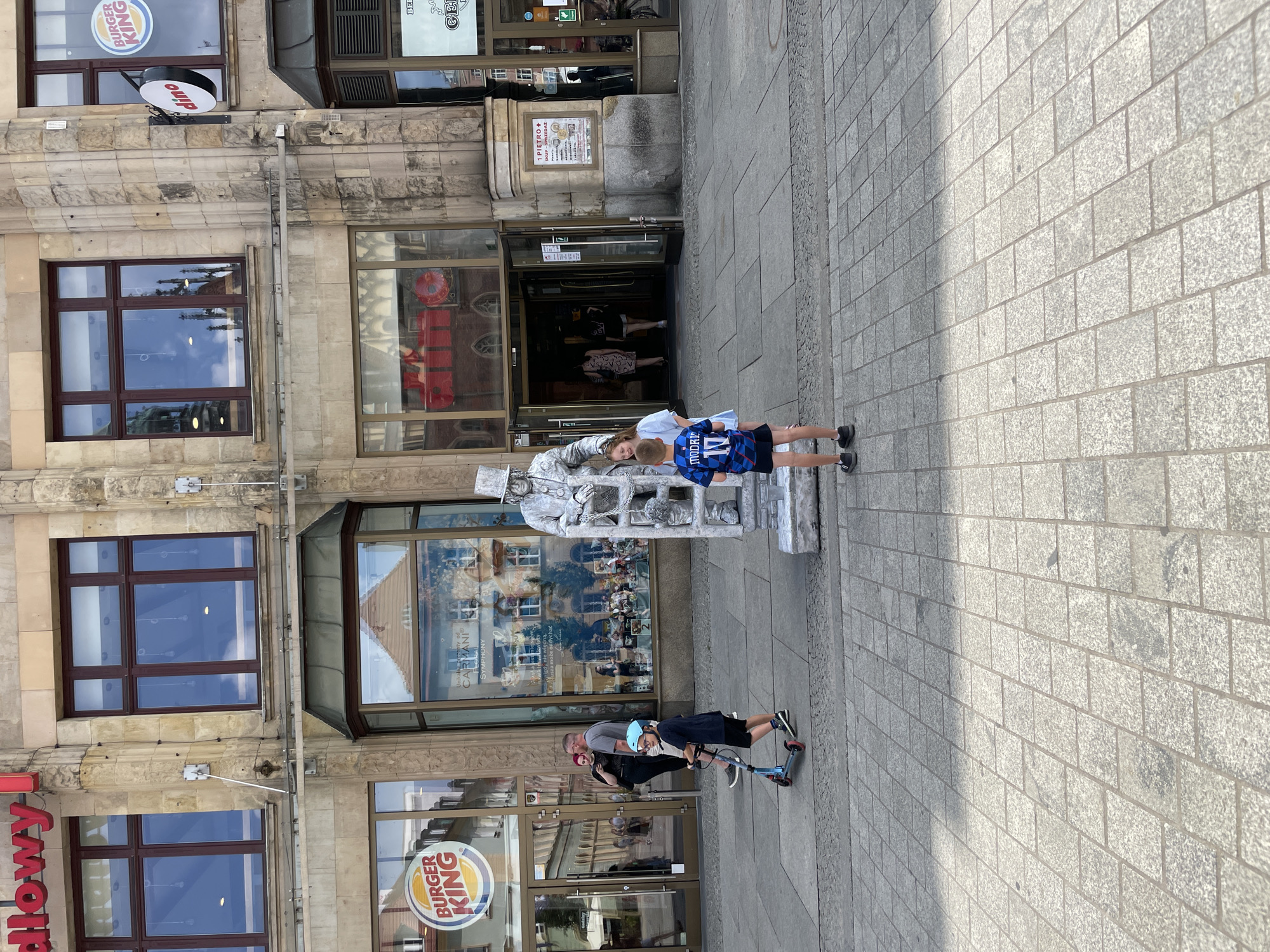 The image depicts a scene on a cobblestone street in front of a building in Wrocław, Poland. On the left, a Burger King restaurant is visible with its logo. A man with short hair and a grey t-shirt, and a woman with dark hair and a black t-shirt are standing near the entrance. A young boy wearing a blue helmet and dark shorts is riding a scooter. In the center of the frame, a street performer dressed as a silver statue, wearing a top hat and holding onto a ladder, is standing on a platform. A young girl with blonde hair and a light blue shirt is standing next to the statue. A young boy wearing a blue and black soccer jersey with the number 17 is standing in the foreground, facing away from the camera. The building facade is made of stone, with large windows. The entrance to a building with glass doors is visible in the background. Other people are seen walking in the background.
