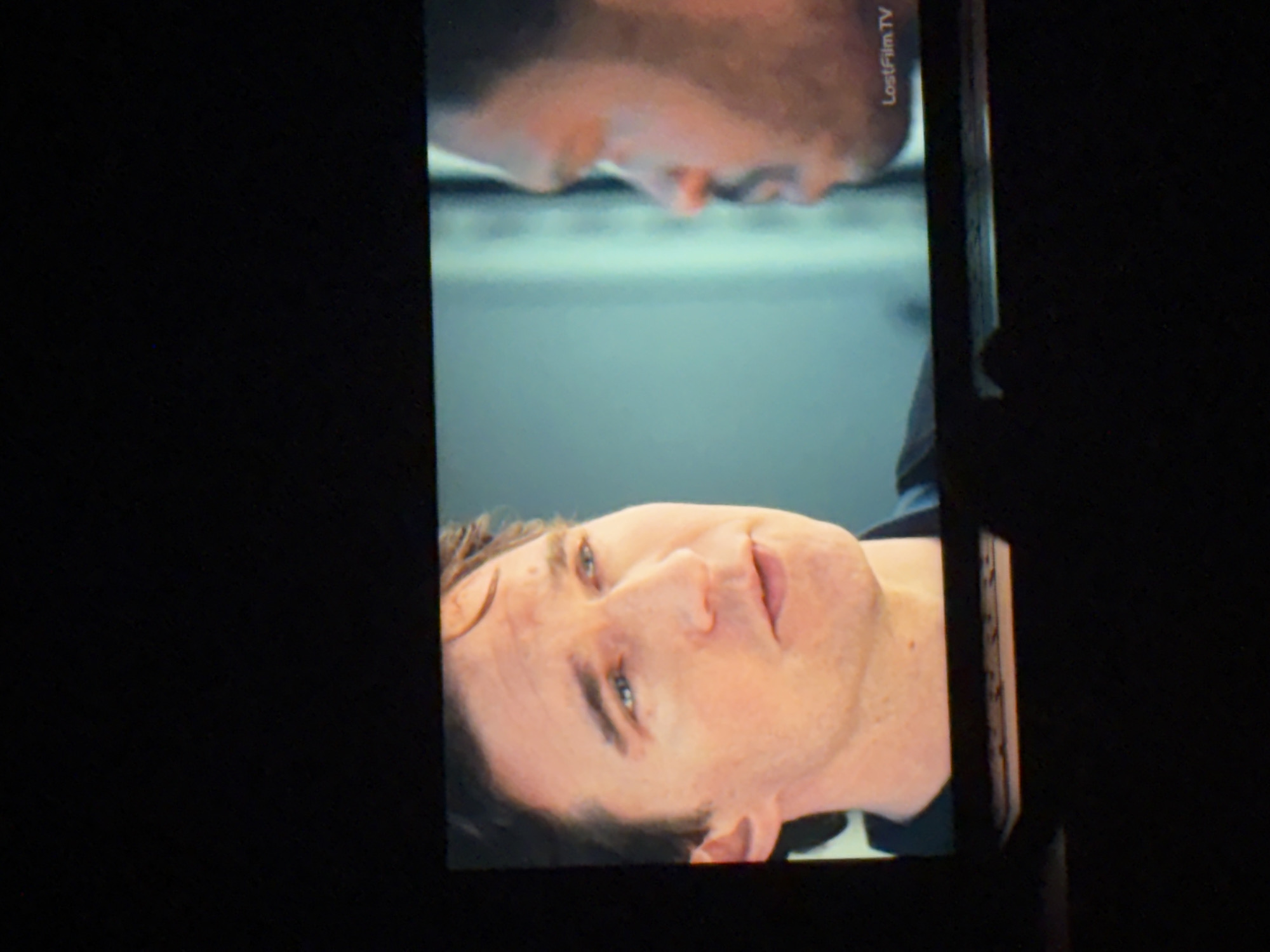 A photograph shows a television screen displaying two men's faces. One face is oriented upright, while the other is inverted above it. The screen is identified with the watermark "LostFilm TV." The image appears to be taken in a dark environment, with the screen's glow illuminating the subjects. There is no evidence of the location being in Budva, Montenegro.