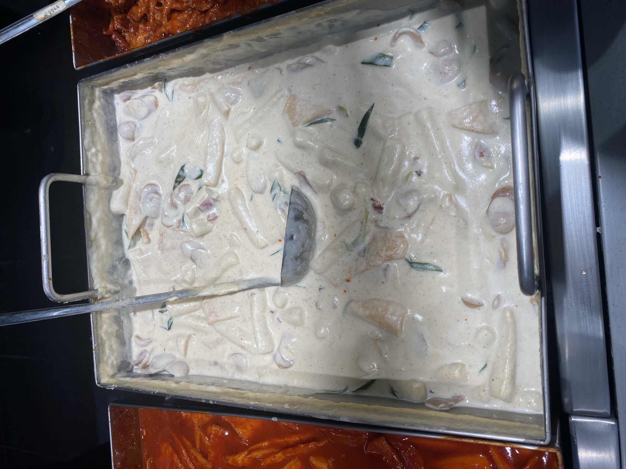 The image shows a close-up, overhead view of two food pans in a buffet-style establishment, likely in North Jakarta, Indonesia. The pan on the left contains a bright red, saucy dish with what appear to be strips of food, possibly tteokbokki. The larger pan in the center is filled with a creamy, off-white sauce containing various ingredients such as rice cakes (tteok), possibly sausages, and green herbs. A metal ladle is partially submerged in this creamy dish. The pans are set into a stainless steel counter, and the background is dark. There are no people visible in the image.