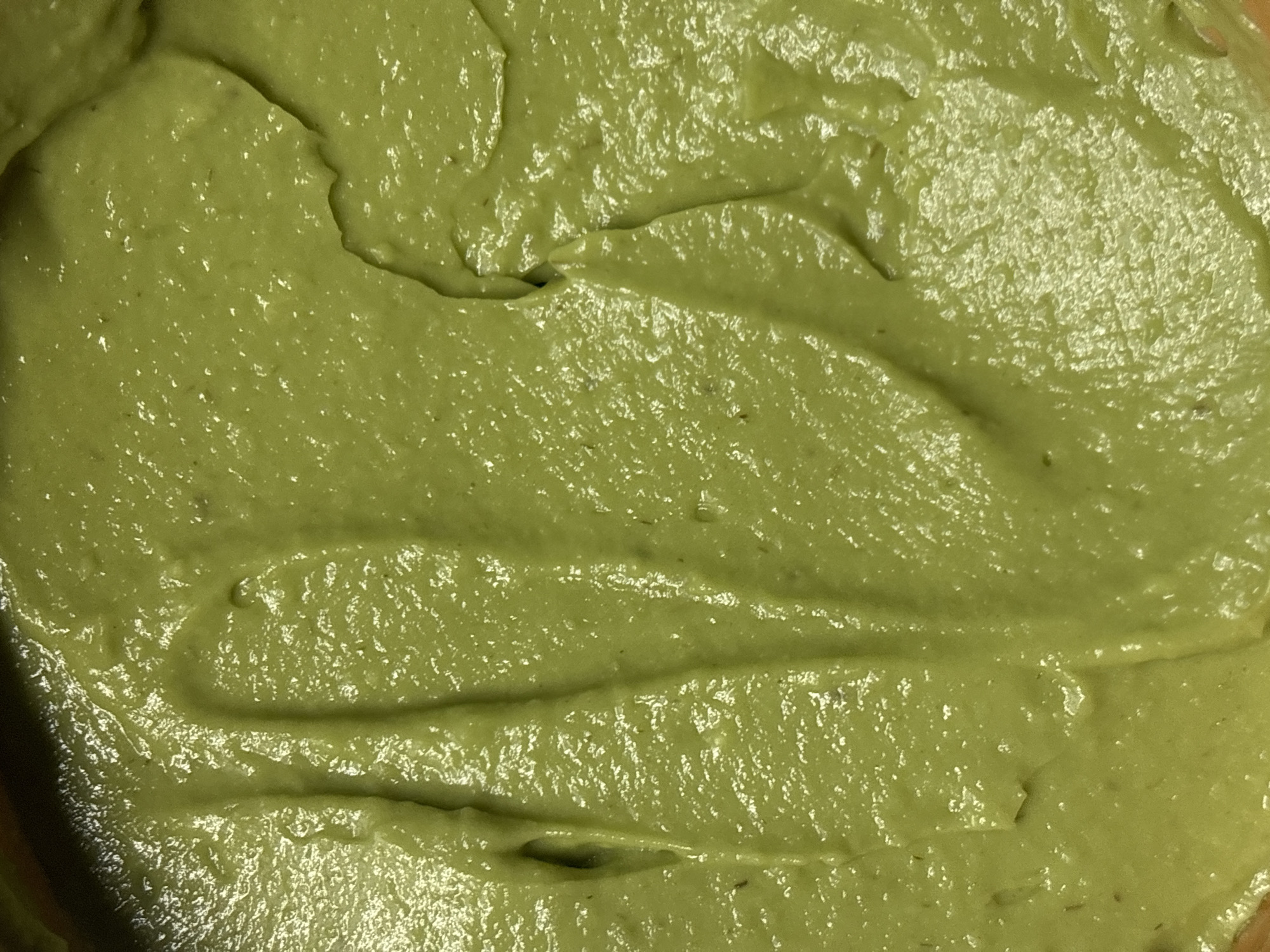 The image displays a close-up, top-down view of a thick, creamy, green substance, most likely a food item such as guacamole or an avocado-based dip or spread. The entire frame is filled with this substance, highlighting its rich, muted olive-green color, which exhibits variations in shade due to light reflections and shadowed contours.
The surface of the substance is characterized by soft, undulating ridges, swirls, and peaks, indicating that it has been recently stirred, spread, or manipulated. A noticeable sheen or glisten is visible across its surface, suggesting a moist or slightly oily consistency. Tiny, dark flecks are embedded within the green matrix, possibly fragments of herbs, spices, or small pieces of avocado, adding to its natural appearance.
Faint edges of what appears to be a light-colored bowl or container are visible along the perimeter of the food, suggesting it is contained and prepared for consumption. No specific activity is taking place; the image captures the static state of the prepared food item. There are no people, discernible objects other than the food and its container, visible text, or specific location cues within the frame to indicate a particular setting, time of day, or weather conditions. The focus is entirely on the textural and chromatic details of the food.