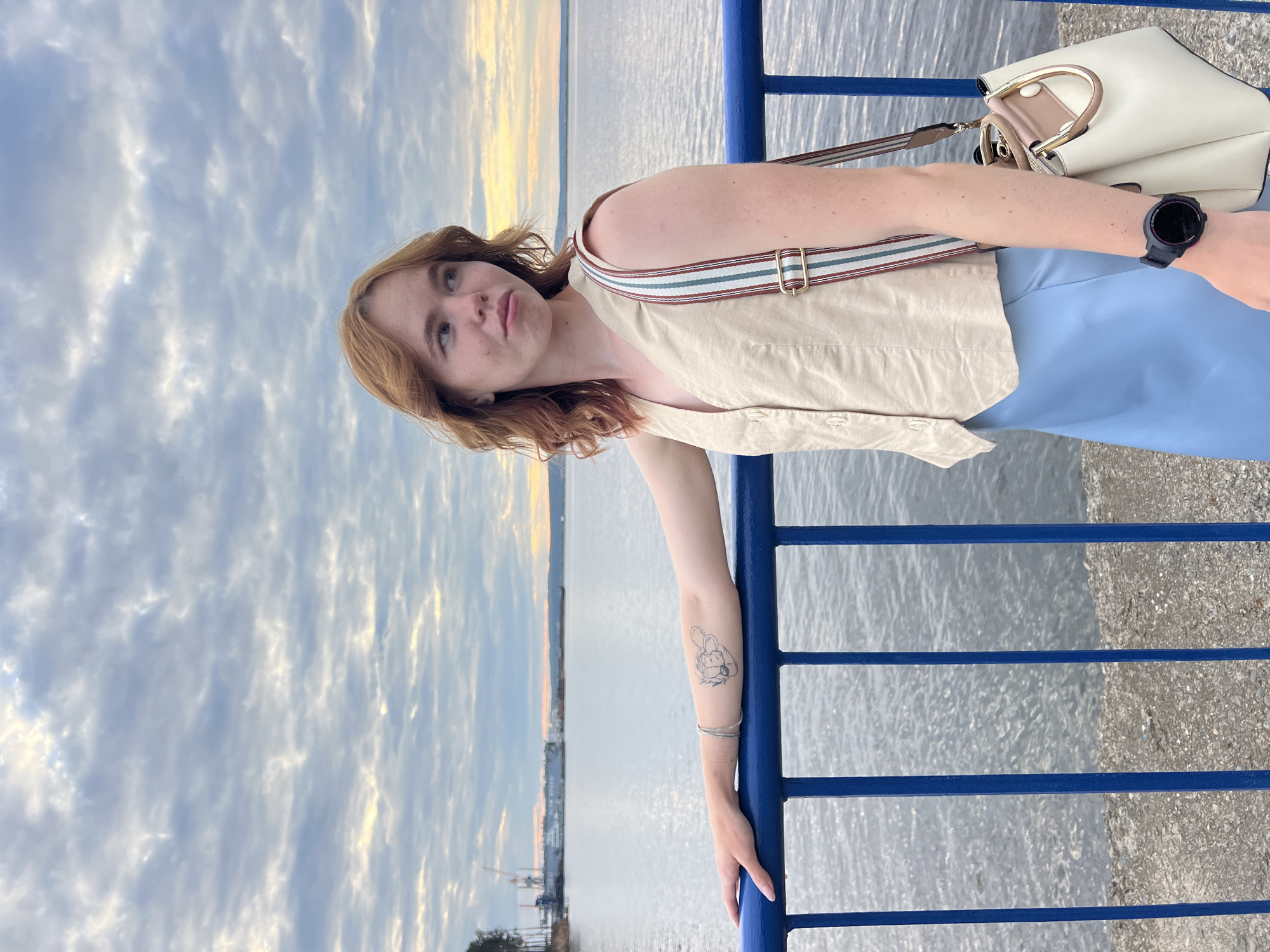 A young woman with reddish-brown hair stands on a walkway, leaning on a blue railing. She is wearing a light beige sleeveless vest over a light blue skirt. A white handbag with a striped strap hangs from her shoulder. She has a tattoo of a cartoon character on her left forearm. In the background, a wide body of water stretches to the horizon, with industrial cranes and structures visible on the far bank. The sky is filled with clouds, with hints of yellow and orange light suggesting either sunrise or sunset. The location is the embankment in the city of Chaikovsky, Russia.