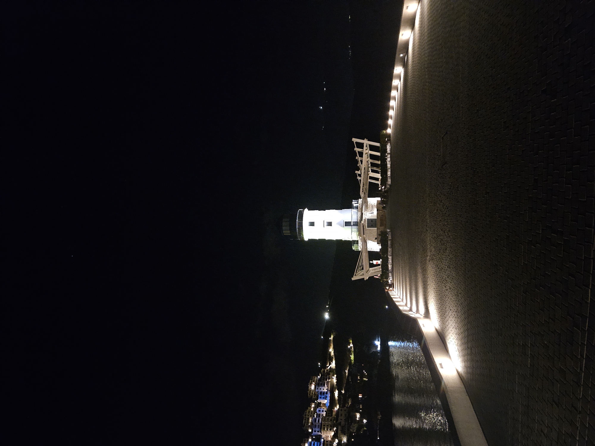 An outdoor, ground-level, wide shot captures a lighthouse and surrounding structures at night. The lighthouse, a tall, white cylindrical structure with black accents, stands prominently in the center-right of the frame. It is illuminated with bright white lights, highlighting its architectural features and the glass surrounding its lantern room.
In front of the lighthouse, a white, modern, open-air structure extends outwards, resembling a cantilevered deck or stage with a slatted roof. This structure is also illuminated.
To the left of the lighthouse, across a body of water, is a hillside covered with buildings. These buildings are multi-story, with white and blue facades, and are lit with various colored lights, creating a vibrant, illuminated village scene. The water reflects the lights from the buildings and the sky.
The foreground of the image is a paved walkway made of dark cobblestones. The walkway is lined with a low, white wall on the left, which is illuminated by a series of evenly spaced, upward-facing lights, casting a warm glow on the paving stones. The walkway curves gently away from the viewer towards the lighthouse.
The sky is a deep, dark black, with no visible stars. The overall atmosphere is one of a well-lit, possibly resort or tourist area, at night. The image is captured in portrait orientation.