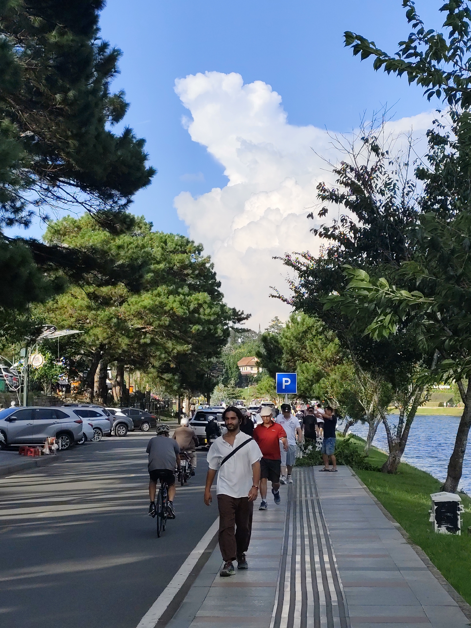 In the city of Xuân Hương, Đà Lạt, Vietnam, a wide, paved road runs alongside a body of water, likely Xuân Hương Lake. Lush green trees line the right side of the road, casting shadows across the asphalt. A concrete sidewalk with tactile paving runs parallel to the road, bordered by manicured grass. Several cars are parked along the left side of the road, and a cyclist in a grey shirt and black shorts rides away from the camera. To the right of the road, a group of people are walking along the sidewalk. One man in a white shirt and brown pants walks towards the camera, looking to his right. Another man in a red shirt and black shorts walks in the same direction. Behind them, a few other individuals are seen walking, and a blue parking sign is visible. The sky is a bright blue, with large, white cumulus clouds dominating the upper portion of the frame.