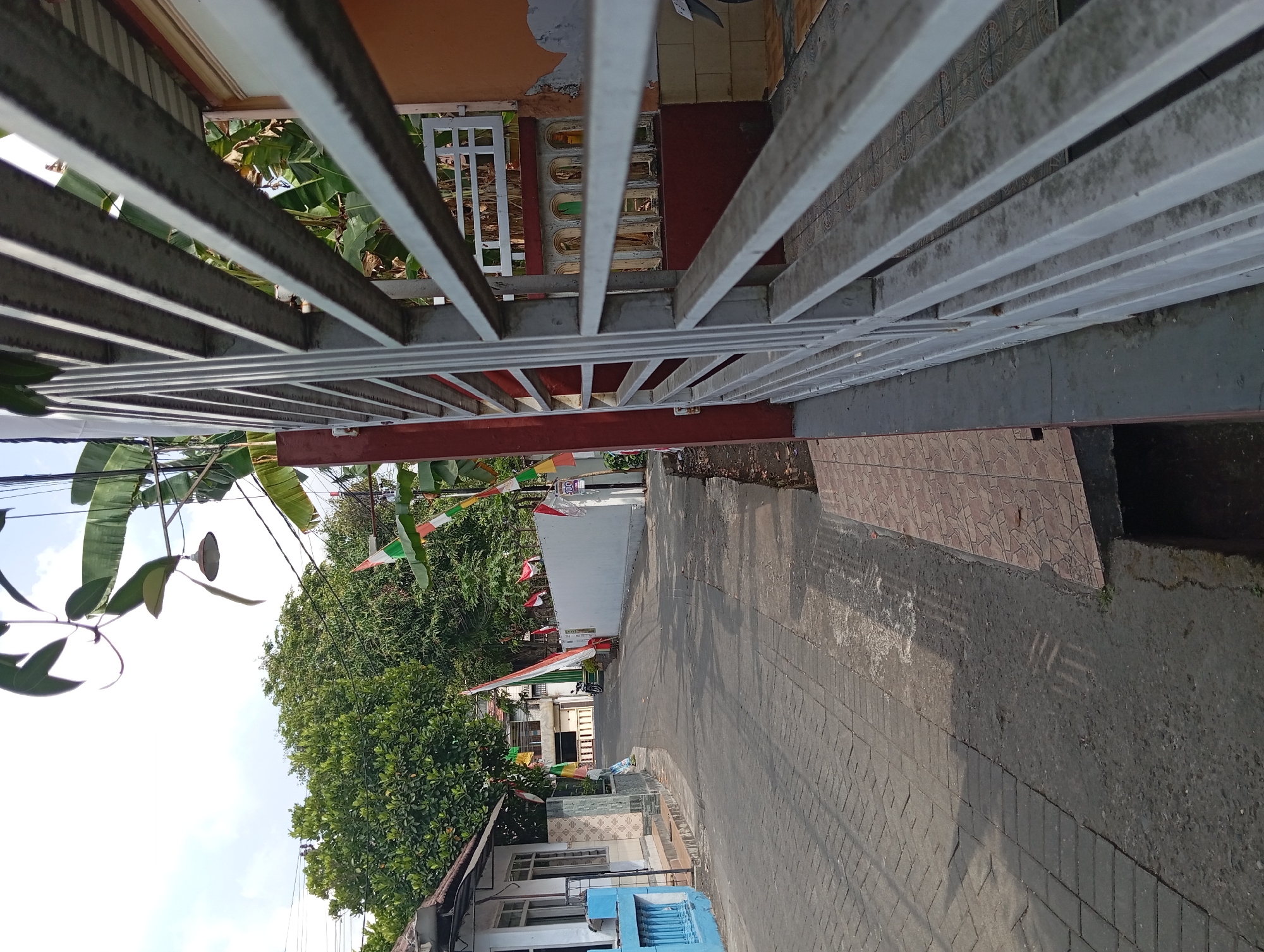 The image depicts a street scene in Kepulungan, Indonesia, on a sunny day. The street is paved with cobblestones on the left side and asphalt on the right. Buildings line the street, with one on the left featuring a white facade and a bright blue railing. Flags, seemingly Indonesian, are strung across the street and along a white wall. Lush green trees and banana plants are visible, adding to the tropical atmosphere. A white metal fence or gate occupies the foreground on the right, obscuring part of the view into a property. The sky is clear with a few wispy clouds. The overall impression is of a quiet residential street, possibly decorated for a celebration.