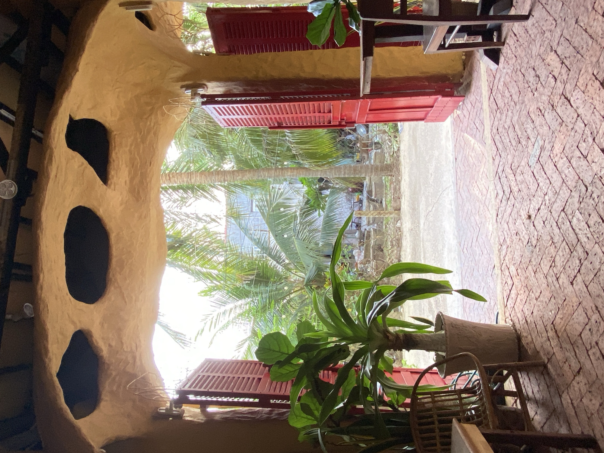 The image shows an outdoor setting in Mũi Né, Vietnam. The scene is a cozy, rustic area with textured, earthy-toned walls and a brick floor. In the foreground, a large potted plant with lush green leaves sits next to a woven chair. Behind the plant, there are open red shutters revealing palm trees and a bright sky, suggesting it's daytime. The architecture appears somewhat organic, with curved walls and natural-looking openings. The overall impression is one of a relaxed, tropical environment, possibly a cafe or guesthouse. The light suggests it could be morning or afternoon, with bright, diffused sunlight. There is no visible text in the image.