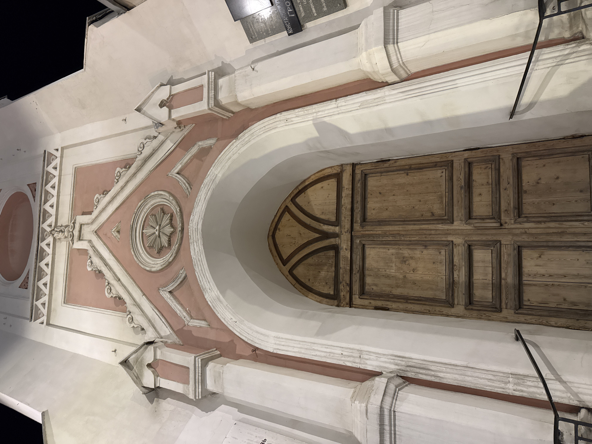 The image depicts the façade of a building, likely a church or a similar historical structure. The building's architectural style features a large archway with a wooden door. Above the arch, there's an ornate triangular pediment with decorative elements, including a circular medallion. The color scheme is predominantly white and pink, with the wooden door providing a contrasting element. A sign with Cyrillic script is visible on the building. The image's angle suggests that it was taken from a low position, looking upwards. The setting is nighttime, indicated by the darkness in the upper left corner. The location appears to be a city, though the specific city and country are not identifiable from the image.