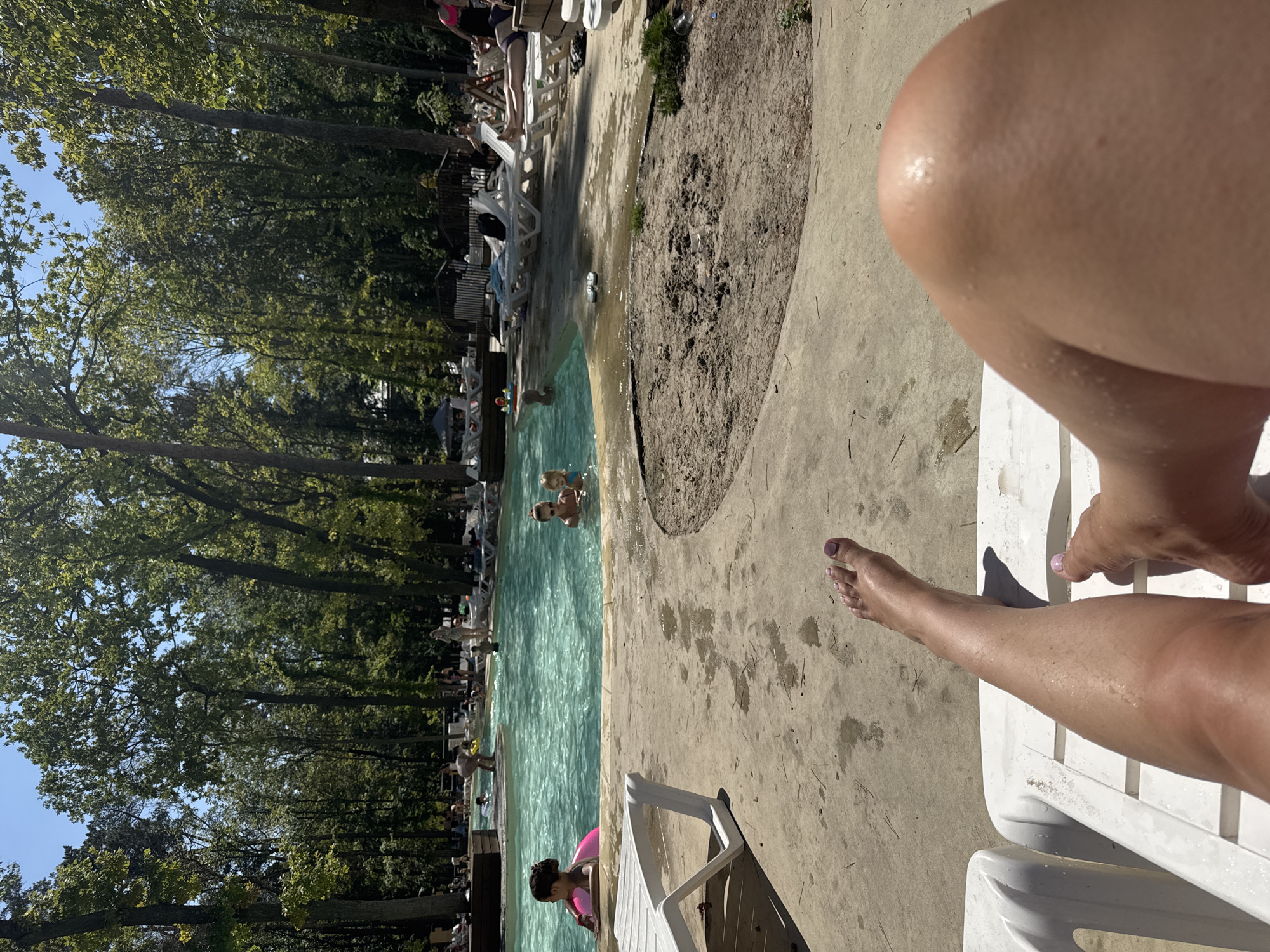 The image depicts an outdoor swimming pool area on a sunny day. The foreground shows a person's legs and feet resting on a white lounge chair. The background features a large swimming pool with several people in and around it, including children and adults. Trees surround the pool area, creating a shaded environment. Some lounge chairs and umbrellas are visible near the pool. The overall scene suggests a leisure activity at a resort or recreational facility. The location is identified as Orlivshchyna, Ukraine.