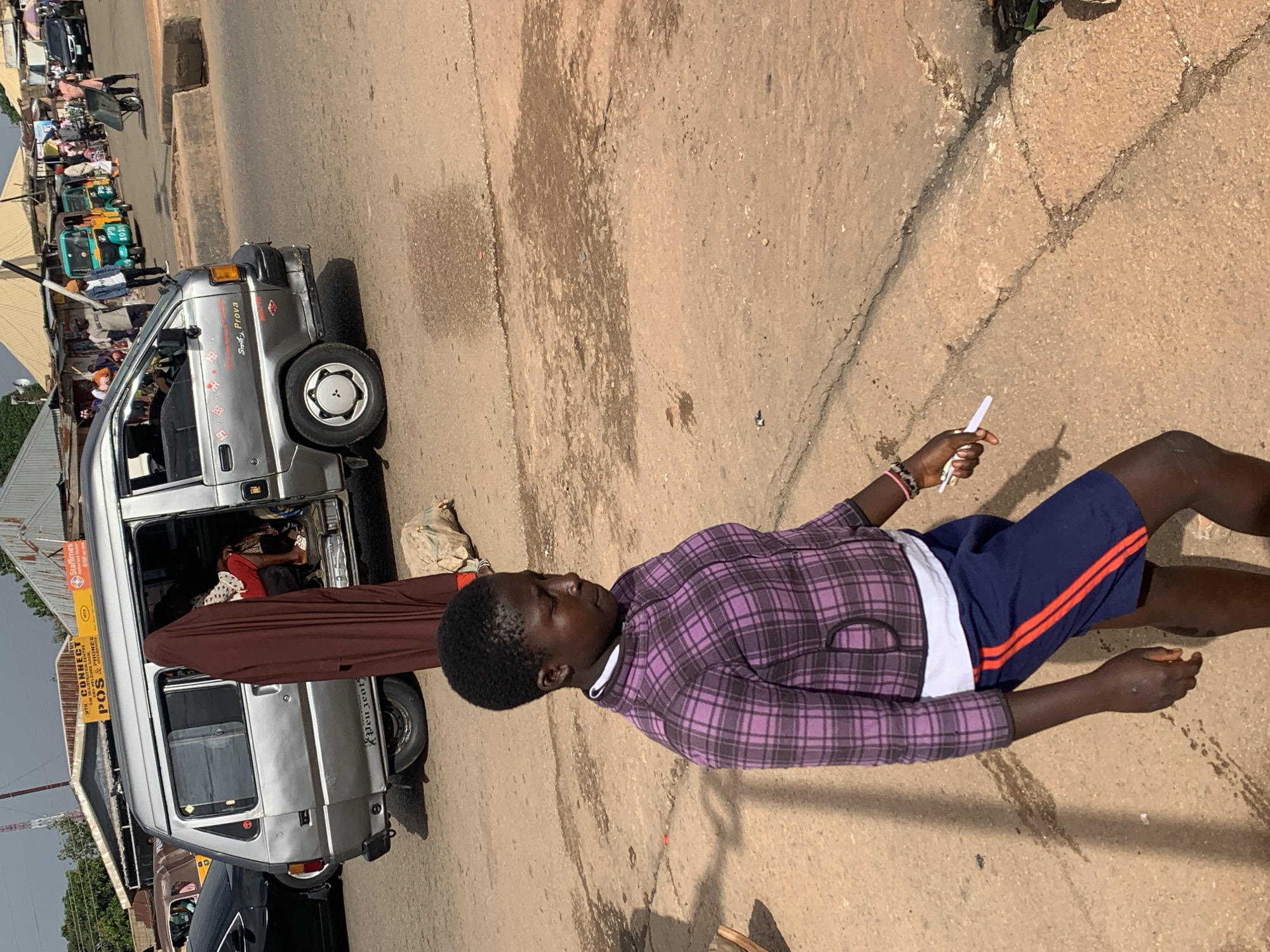 In Gura Top, Nigeria, a young person walks on a dirt road next to a silver minivan. The individual is wearing a purple plaid jacket, a white shirt, and navy shorts with red stripes. They are holding a white object, possibly a cigarette, in their right hand. The minivan has tinted windows and a "MTN CONNECT" sign on top, indicating it's likely a commercial vehicle. In the background, several other vehicles, including colorful rickshaws, are visible, along with buildings that appear to be shops or stalls. The overall scene suggests a bustling street in a Nigerian town.