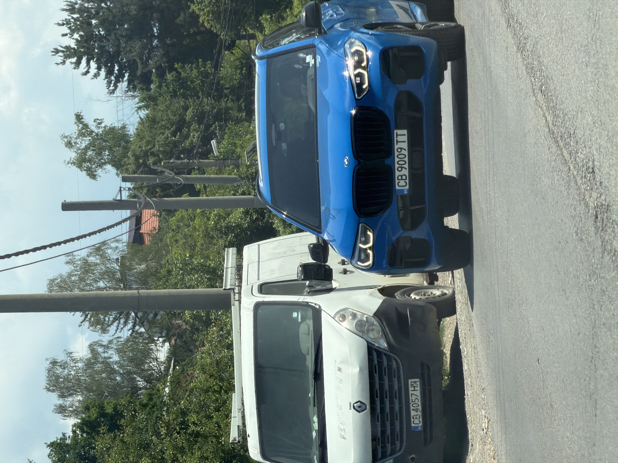 In Sofia, Bulgaria, on a roadside, a blue BMW X5 is parked next to a white Renault van. The BMW, with license plate CB 9009 TT, is in the foreground, its headlights on. The van, with license plate CB 4057 HM, is to the left of the BMW. The scene is illuminated by daylight under a cloudy sky. Trees and telephone poles line the background.