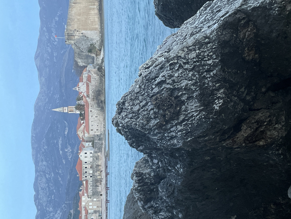 The image displays a coastal scene featuring the Old Town of Budva, Montenegro, viewed across a body of water. In the foreground, large, dark, textured rock formations occupy the lower half and extend towards the right, showing rugged, weathered surfaces.
The midground consists of calm, turquoise-blue water. Beyond the water, the fortified Old Town of Budva rises, characterized by numerous light-colored stone buildings with reddish-orange tiled roofs. A prominent, tall, slender white church bell tower with a pointed spire is visible near the center of the Old Town's skyline. To its right, a robust, high stone fortification wall, likely part of the Citadela, stands at the water's edge, topped with a small red flag, indicative of the Montenegrin national flag. To the left of the bell tower, a lower stone wall fronts additional Old Town structures. A narrow pier or jetty extends into the water from the left side of the Old Town, where two small, indistinct human figures are discernible, walking.
The background features dark, undulating mountain ranges extending across the horizon under an overcast or twilight sky. The overall lighting is subdued, creating muted tones throughout the scene.