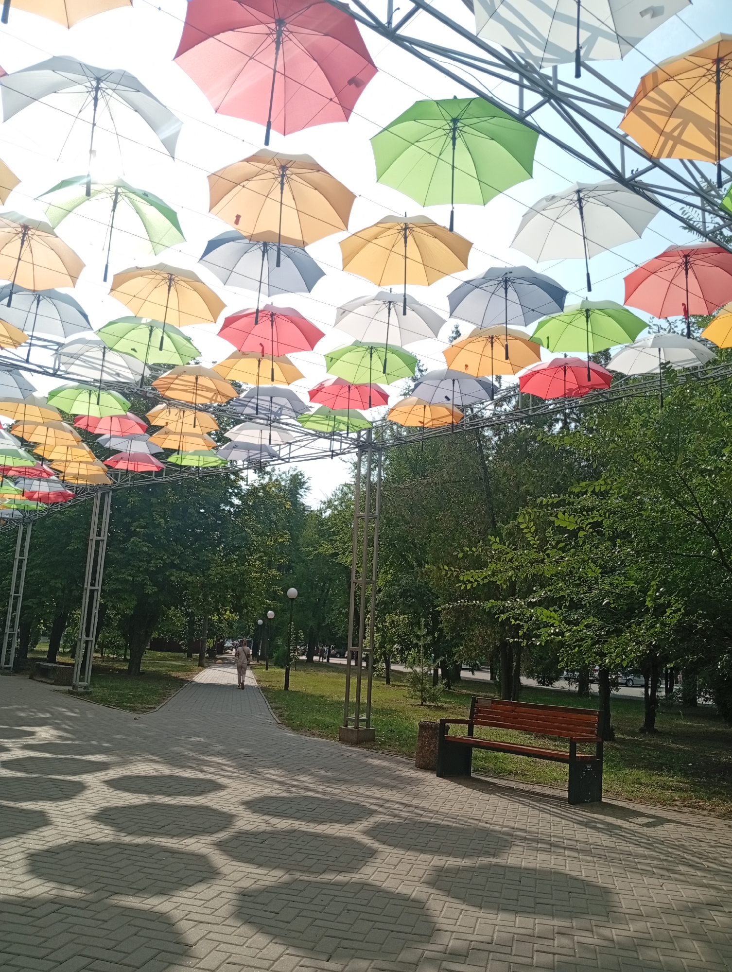 The image depicts a park in the city of Pokrov, Ukraine, adorned with a colorful canopy of umbrellas suspended overhead. The umbrellas are of various colors, including pink, red, orange, yellow, green, and white, creating a vibrant and festive atmosphere. They are strung across a metal framework, casting intricate shadows on the paved walkway below.

In the foreground, a gray brick pathway winds through the park. To the right of the path, a wooden bench with dark metal legs is situated, facing the walkway. A trash receptacle stands next to the bench. The path is illuminated by sunlight, which also casts distinct hexagonal shadows from the umbrellas onto the ground.

A lone individual, a woman with her back to the camera, walks away from the viewer along the path. She is wearing light-colored clothing and carrying a bag over her shoulder. The path leads towards a line of trees that form the edge of the park. Beyond the trees, glimpses of a road with passing cars can be seen, suggesting the park is in a relatively urban setting.

The background is dominated by lush green trees, their leaves rustling in what appears to be a sunny day. The sky above is a clear blue. The overall scene conveys a sense of leisure and recreation within a well-maintained urban park.