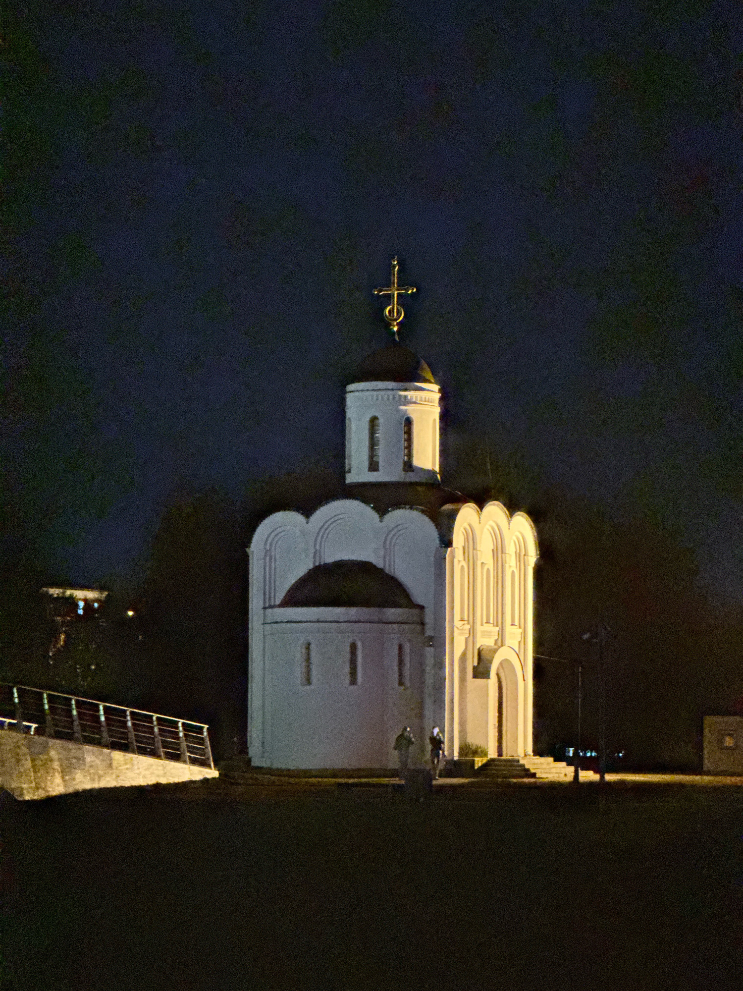 A white, onion-domed church with a golden cross on top is illuminated at night. The church has arched windows and decorative arches above its entrance. To the left of the church, a concrete bridge or walkway with metal railings is lit by artificial lights. Two figures, appearing to be people, are standing near the entrance of the church. In the distance, across a body of water, a building with illuminated windows is visible. The overall scene is dark, with the sky appearing deep blue or black, indicating nighttime. The image is grainy, suggesting it was taken in low light conditions. This church is located in the city of Tver, Russia, and is known as the Church of the Assumption of the Blessed Virgin Mary, situated on the banks of the Volga River.