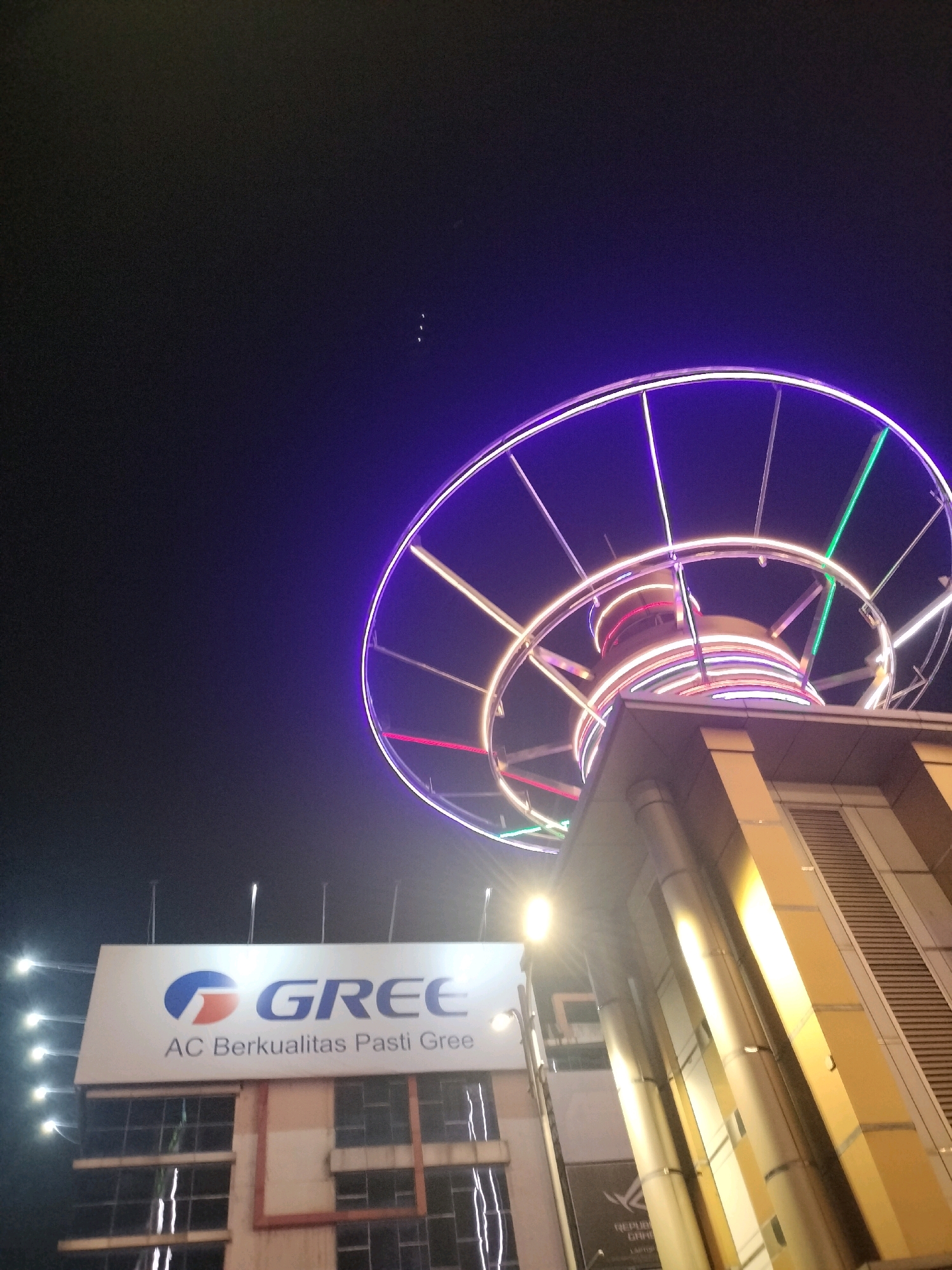 The image captures a night view of a Ferris wheel illuminated with colorful lights, positioned above a building in Batam, Indonesia. The Ferris wheel is the dominant feature, with its circular structure and spokes adorned with vibrant neon lights in shades of purple, blue, green, and red. Below the Ferris wheel, a building is visible, featuring a large sign for "GREE" air conditioners with the tagline "AC Berkualitas Pasti Gree". The building itself appears to be a multi-story structure with windows and architectural elements. The sky above is dark, indicating it is nighttime. Several streetlights are also visible, adding to the ambient lighting of the scene. The overall impression is that of an urban nightscape, likely a commercial or entertainment area in Batam.