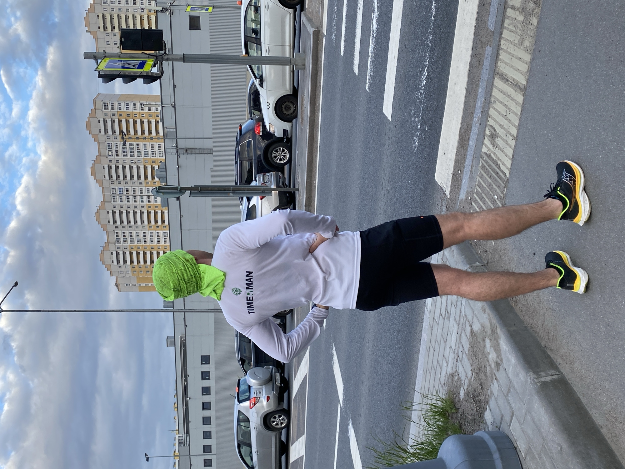 A man stands at a crosswalk on a city street. He wears black shorts, a white long-sleeved shirt with the word "TIME:MAN" on the back, a green head covering, and black and yellow running shoes. The crosswalk is marked with white stripes on dark asphalt. Behind the man, cars are parked parallel to the sidewalk. Tall buildings are visible in the background. The sky is overcast with clouds. The location is Saint Petersburg, Russia.