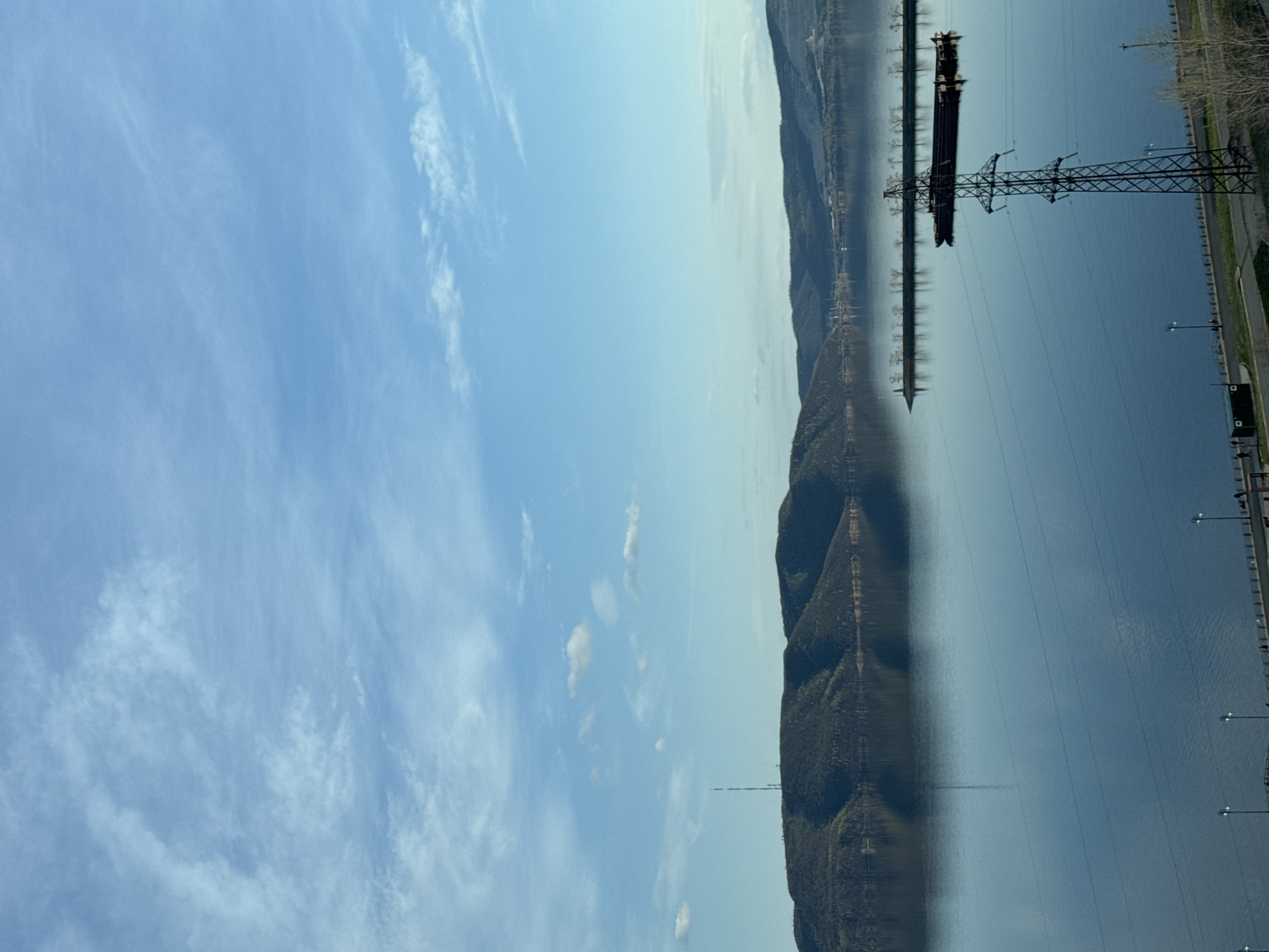 A wide landscape photograph, when correctly oriented, captures a broad body of water, likely a river or large lake, stretching horizontally across the middle of the frame. Above, a vast, light blue sky is adorned with scattered, wispy white clouds. The water is calm and highly reflective, mirroring the sky and the distant land.
Along the far bank of the water, a long range of mountains or large hills is visible, densely covered in dark green and brown vegetation. Their reflections are distinct in the tranquil surface of the water. On the right side of the image, a tall, dark metal electricity pylon or transmission tower stands prominently, supporting multiple power lines that extend across the water.
To the right of the water's edge, there is a paved area or road lined with several streetlights. Behind the transmission tower, a dark, elongated object, possibly a barge or cargo ship, is situated on the water, its form partially obscured by power lines and its own reflection. Faint details at the base of the distant hills suggest the potential outline of a settlement or structures.
The scene takes place on a clear and bright day, likely late morning or early afternoon, indicated by the well-lit landscape and the presence of only light, high-altitude clouds. No people are visible, and there is no discernible text within the image. The overall impression is a blend of natural vastness with elements of human infrastructure.