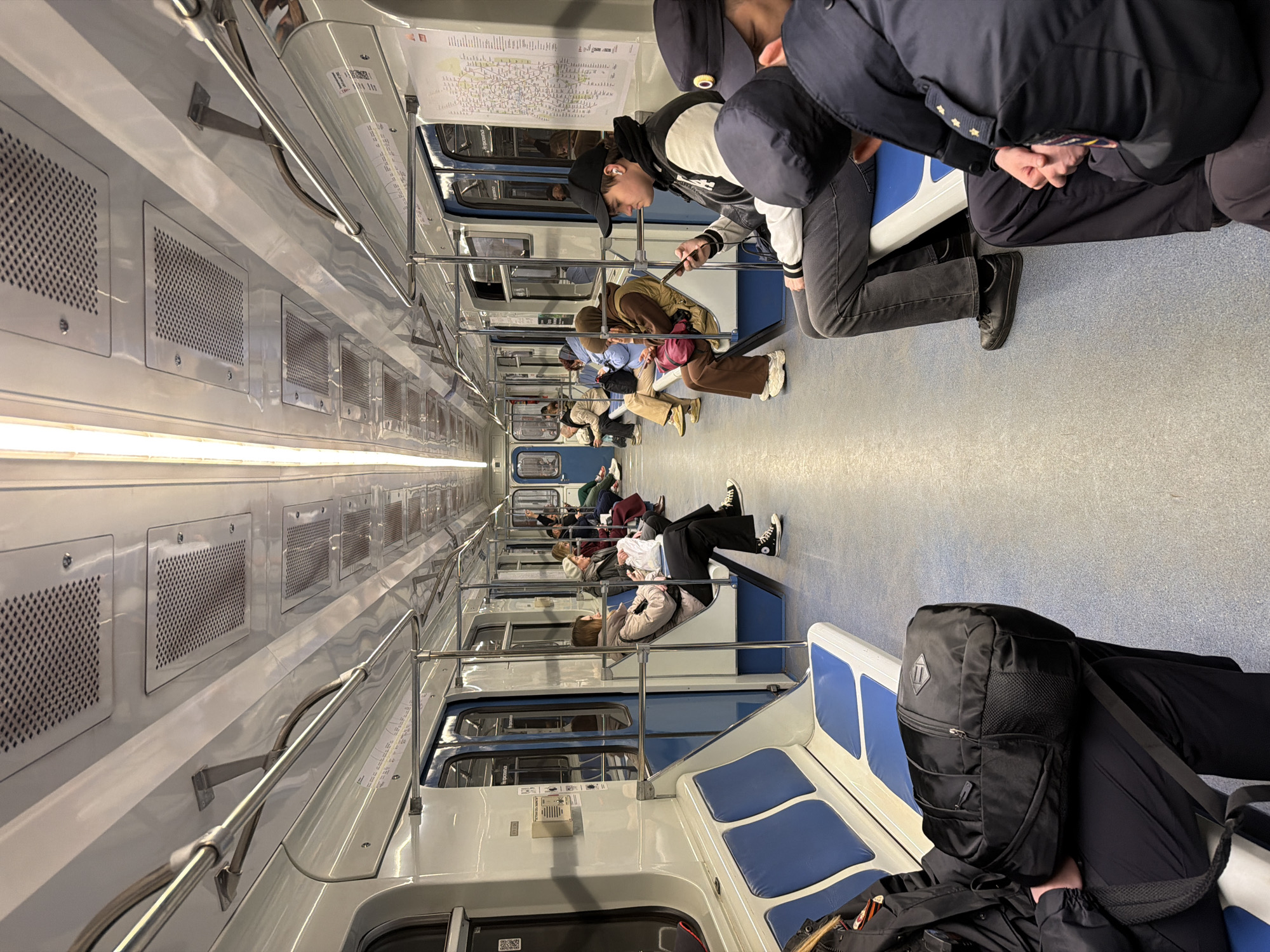 The interior of a subway car with several passengers is shown. The car has white walls with rectangular perforated panels along the upper section. Metal handrails run horizontally along the length of the car. Seats are arranged along the sides, with blue cushions. Passengers are seated, with some looking at their phones. At the front of the car, a map is visible on the wall. There is a black backpack in the lower right corner, obscuring part of the seating area. The city is unknown.