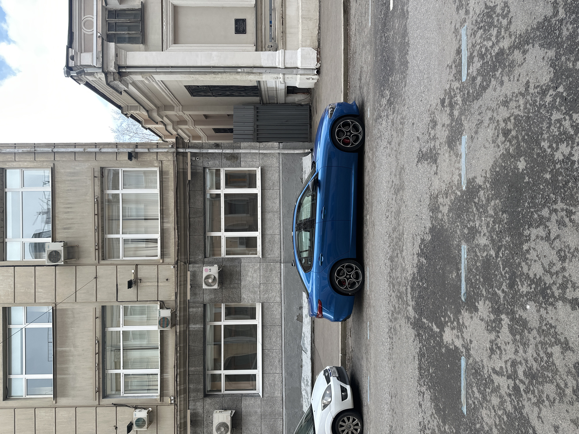 The photograph shows a street scene in Moscow, Russia. A blue car is parked alongside a building with grey facade. The building has multiple windows and air conditioning units mounted on the exterior. A second, white car is partially visible behind the blue one. The street has faded lane markings.