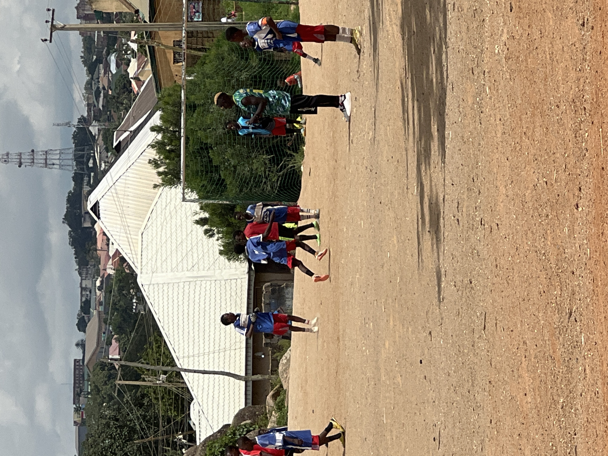 A group of young boys, likely playing soccer, are scattered across a dirt field in Jos, Nigeria. The scene is set against a backdrop of scattered buildings and lush green trees, indicative of an urban or peri-urban environment. One boy, dressed in a green and yellow camouflage jersey, stands out from the others who are mostly in blue and red soccer kits. He appears to be observing the game. Another boy in a blue jersey is seen running. The field itself is uneven and composed of sandy soil, with a soccer goal visible in the background. The sky is overcast with scattered clouds, suggesting a typical weather pattern for the region. The overall impression is of a casual soccer match taking place in a communal outdoor space within the city of Jos.