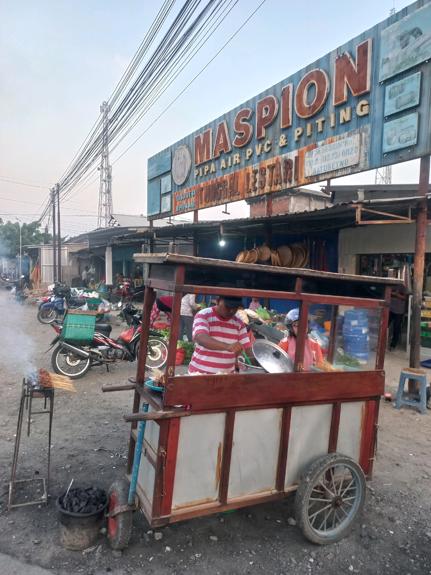 This image depicts an outdoor scene in Baturetno, Indonesia. In the foreground, a food cart is positioned on a dirt road, with a man in a striped shirt actively cooking. The cart has wooden framing and metal siding, and is equipped with wheels. Adjacent to the cart, a small grill with skewers of meat is seen emitting smoke, and a bucket filled with charcoal. Several motorcycles are parked nearby, some with baskets. In the background, a row of shops with awnings lines the street, and a large sign with the text "MASPION PIPA AIR, PVC & PITING" is visible above the shops. The sky is overcast, and power lines and a telecommunications tower are present in the distance. The overall environment suggests a bustling market or roadside commercial area.