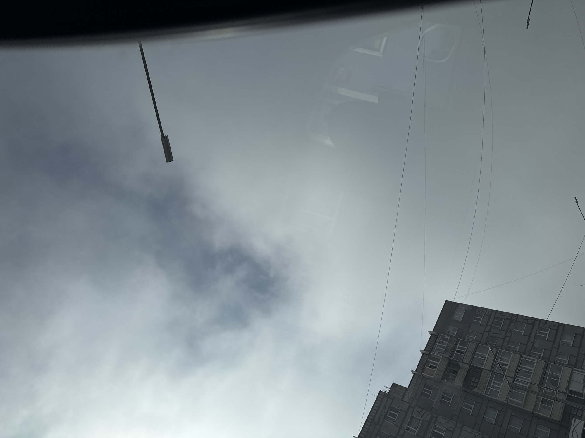 The scene depicts an urban environment in Magadan, Russia, viewed through a reflective surface, likely a vehicle window. The sky is overcast with a gradient of grey clouds, featuring a brighter area in the upper left and a more defined, darker cloud mass in the upper center.
In the lower-left, a multi-story, grey residential building with numerous windows and balconies rises diagonally. Its facade displays vertical dark lines and patterns consistent with aging or structural repairs. Multiple thin utility wires crisscross the sky, extending from the left towards the right. A single streetlight fixture, comprising a long horizontal arm and a rectangular LED lamp, projects into the upper-right sky.
Prominent reflections are visible across the middle and right portions of the image. These reflections include an indistinct interior form resembling a vehicle's window frame and a dark shape, possibly a human head or shoulder, within the upper-right reflected area. A partial reflection of a second streetlight fixture is present in the lower-right corner.