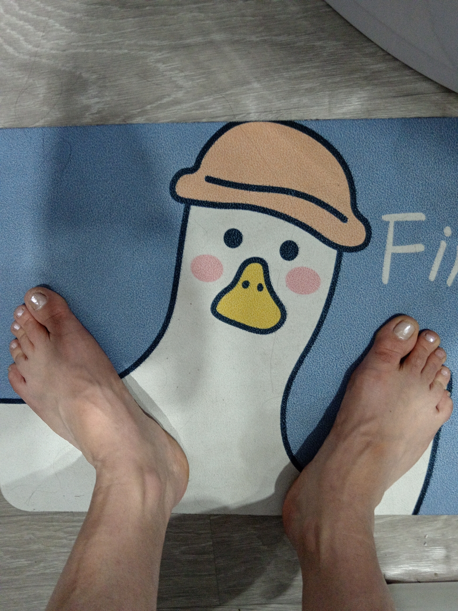 The image displays a close-up, top-down view of two bare feet with painted toenails positioned on a light blue mat featuring a cartoon duck wearing a hat. The mat is placed on a gray wooden floor. The left foot is positioned on the left side of the mat, and the right foot is on the right side. The duck graphic is in the center of the mat, with its head and upper body visible. The background consists of the wooden floor and a corner of a white object, possibly a wall or furniture, at the bottom right. The lighting suggests an indoor setting. The location is not discernible from the image.