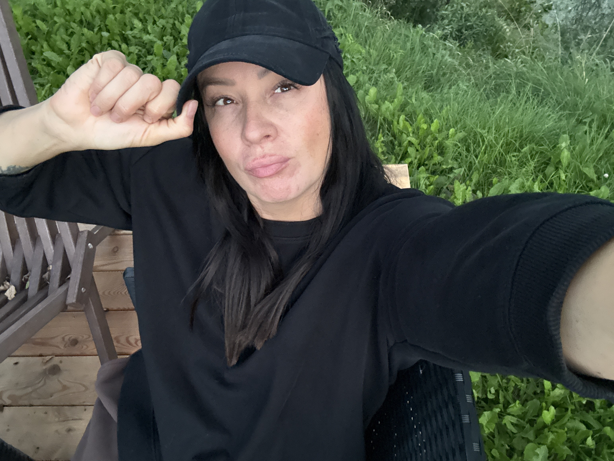 A woman is lying on her side, looking at the camera. She is wearing a black baseball cap and a black sweatshirt. Her hair is dark and straight, and falls over her shoulders. Her skin is fair, with freckles and some acne scarring visible on her face. She has light brown eyes and full lips. Her right arm is bent, with her fist resting near her head, and her left hand is holding the brim of her cap.
The woman is positioned on a wooden deck, with a blurred background of green grass and some foliage. There appears to be a piece of wooden furniture, possibly a chair or bench, partially visible behind her. The lighting suggests it is daytime, possibly overcast, given the soft shadows. The overall scene appears to be outdoors, in a natural setting, possibly a backyard or park.
There are no specific visual cues within the image that can definitively place this scene in the city of Chaikovsky, Russia.