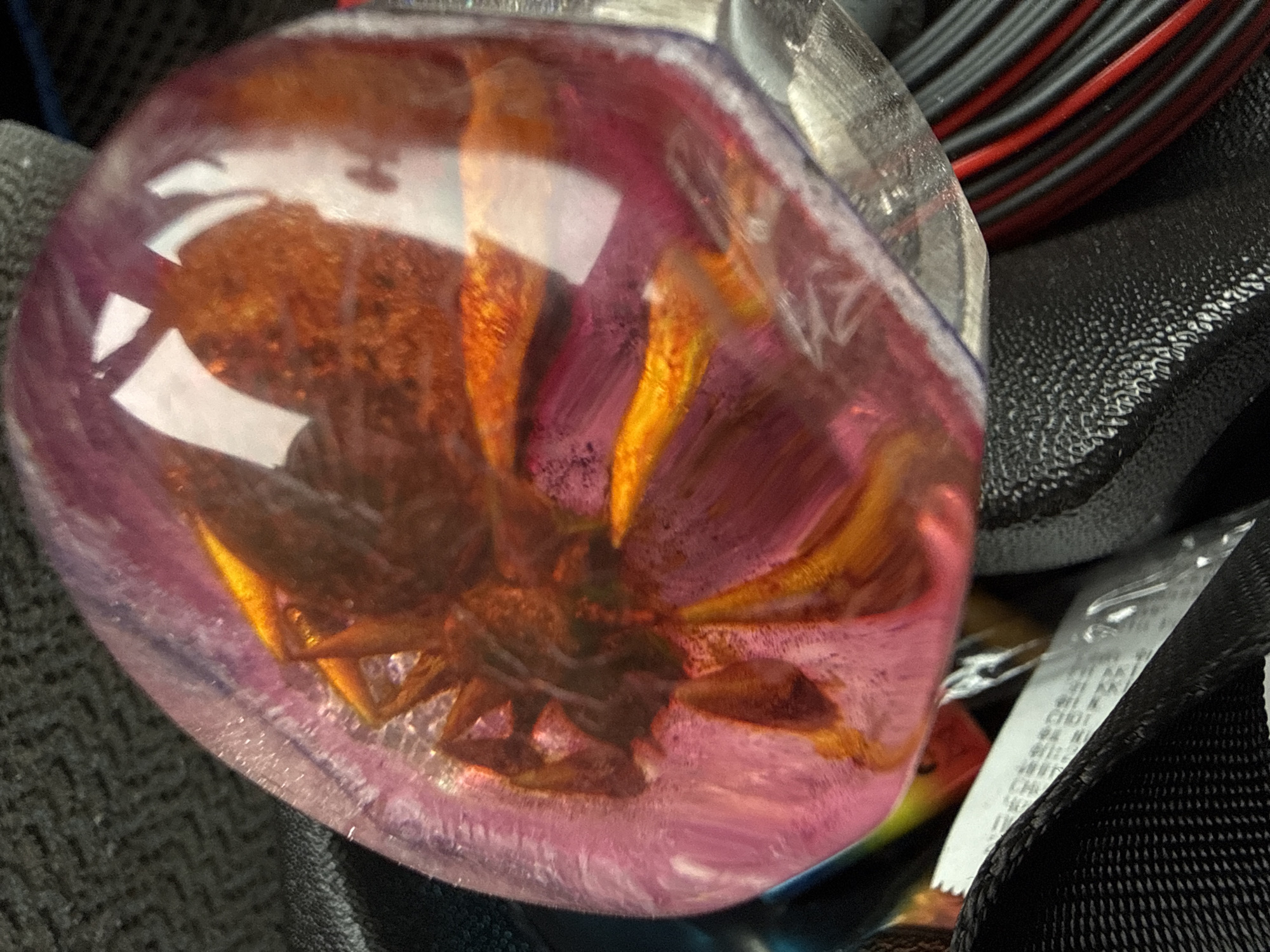 The image depicts a spider encased in clear, pink-tinted resin. The spider is brown and has prominent legs. The resin is clear but has a pink hue to it and contains white specks. The resin object is surrounded by various objects, including black textured fabric, black and red striped cables, and a partially visible white label with text. The scene is dimly lit. There is no explicit evidence to indicate that the picture was taken in Chaikovsky, Russia.