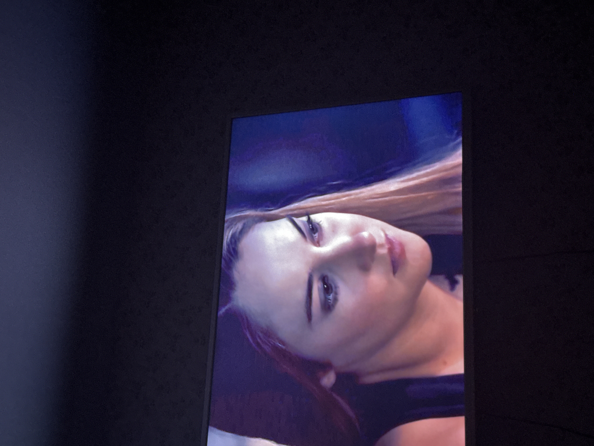 A close-up photograph shows a television screen displaying a woman's face. The woman has fair skin, light brown hair, and dark eyebrows. She is wearing a black top. The background of the image on the screen appears to be a blend of blue and purple hues. The television screen is positioned against a dark wall, possibly in an indoor setting. The location is in Mytishchi, Russia, as that was specified.