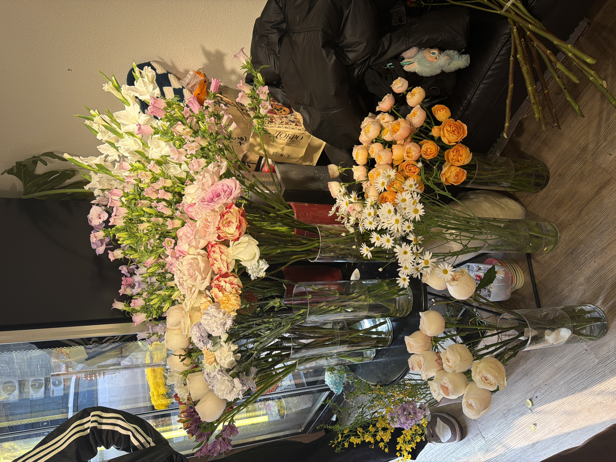 The image depicts an indoor scene with an assortment of flowers in vases. These include gladioli, lisianthus, roses, carnations, daisies, and other unidentified blossoms. They are predominantly pink, white, orange, and yellow. The vases appear to be arranged on a surface near a window, reflected in which is an exterior scene. A black puffer jacket, a shopping bag marked with Cyrillic script, a knitted object, and bare flower stems are also visible. The setting implies the presence of a florist's workspace or flower arranging area. There is no verifiable information to suggest that the image was taken in Jiangning District, China.