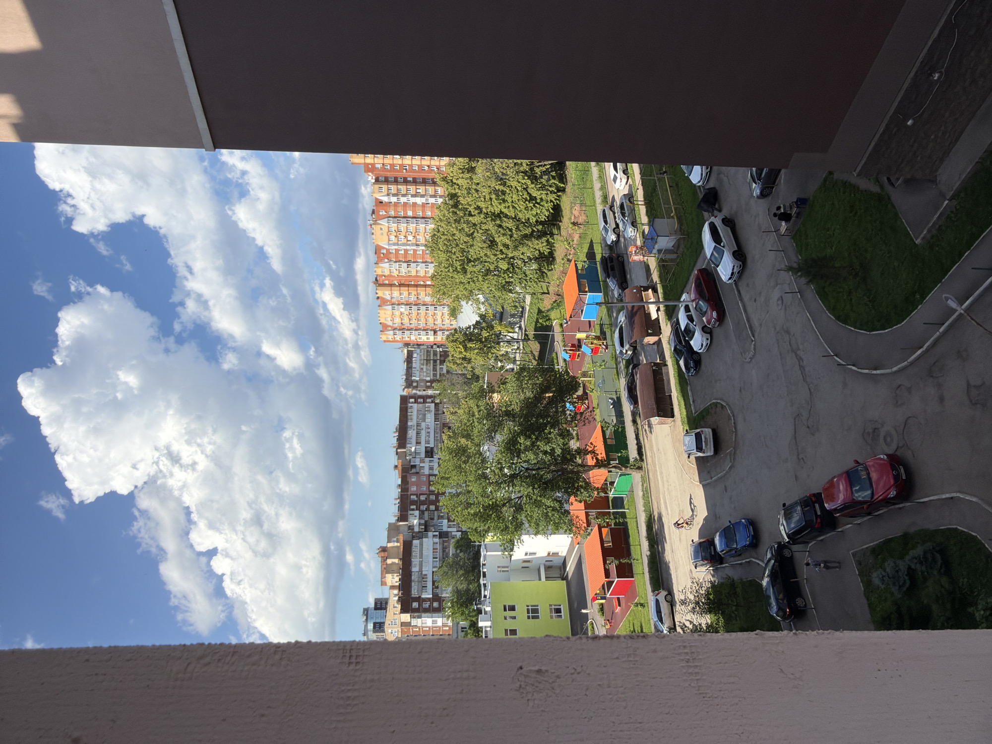 This image depicts a view from a building overlooking a residential courtyard in Ufa, Russia. The scene is dominated by a bright blue sky with fluffy white clouds. In the foreground, a textured, light-colored wall from the building where the photo was taken partially obstructs the view on the left. To the right, a dark brown building facade runs vertically.
The courtyard below features a playground with colorful structures, including orange and red shelters and blue play equipment. Several trees, some with lush green foliage, are scattered throughout the area. A paved road or parking lot occupies a significant portion of the courtyard, with numerous parked cars of various colors, including black, white, blue, and red. Some cars appear to be in a line, suggesting a parking arrangement.
In the background, multiple apartment buildings of varying heights and architectural styles are visible. One prominent building is a multi-story structure with a facade featuring orange, white, and brick-colored sections. Other buildings in the distance appear to be older, with a more uniform brick design.
A few individuals are present in the courtyard. One person is standing near a black car, and another is seen sitting on a bench with another individual. A person on a bicycle is riding on the paved area. The overall impression is a typical urban residential setting on a sunny day.