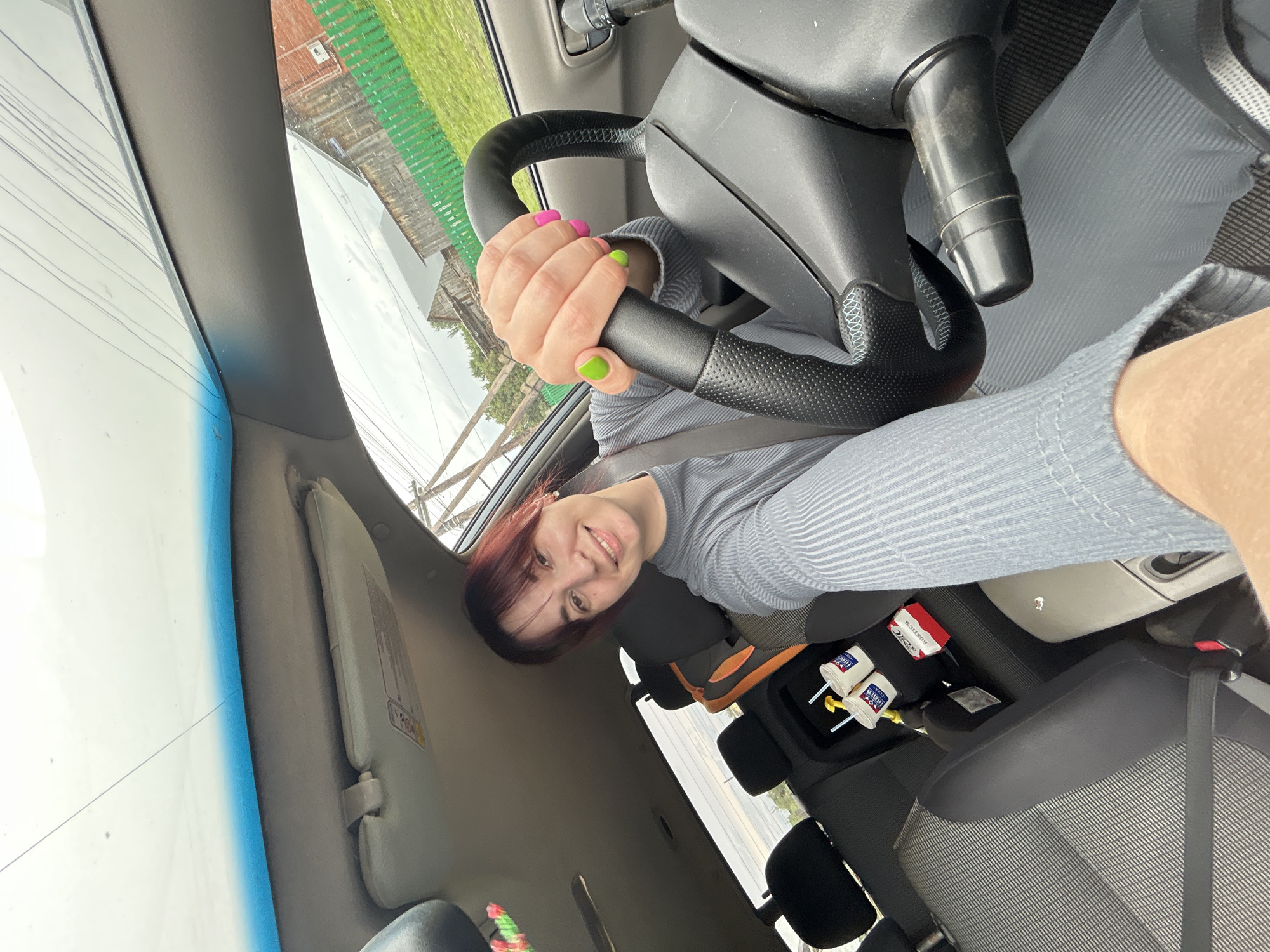 In the city of Fokino, Russia, a young woman with dark red hair, wearing a gray ribbed sweater, is captured in a selfie from the driver's seat of a car. She is smiling and holding the steering wheel, her hands adorned with bright pink and lime green nail polish. The car's interior is visible, with a child car seat in the back and two cups with straws placed in a holder. Outside the car, a wooden fence and building can be seen, suggesting a rural or semi-rural setting. The sky is overcast, indicating cloudy weather.