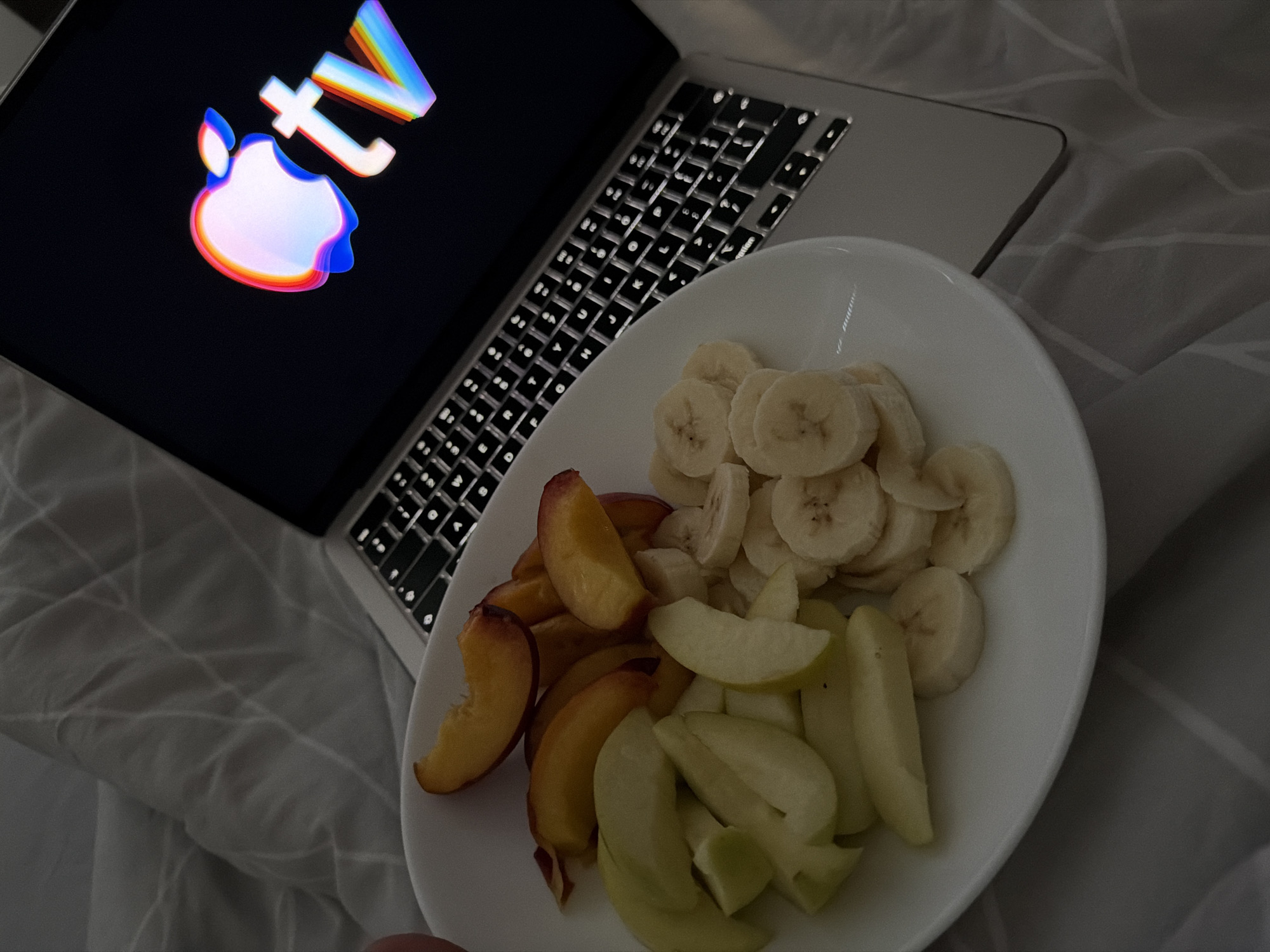 The image shows a white plate filled with sliced fruits, including peaches, apples, and bananas, placed on top of a laptop keyboard. The laptop screen displays the Apple TV logo with a colorful, distorted effect. The scene appears to be set in a bedroom, with a grey, patterned duvet or blanket visible in the background. No people are present in the frame. The location is not specified in the image.
