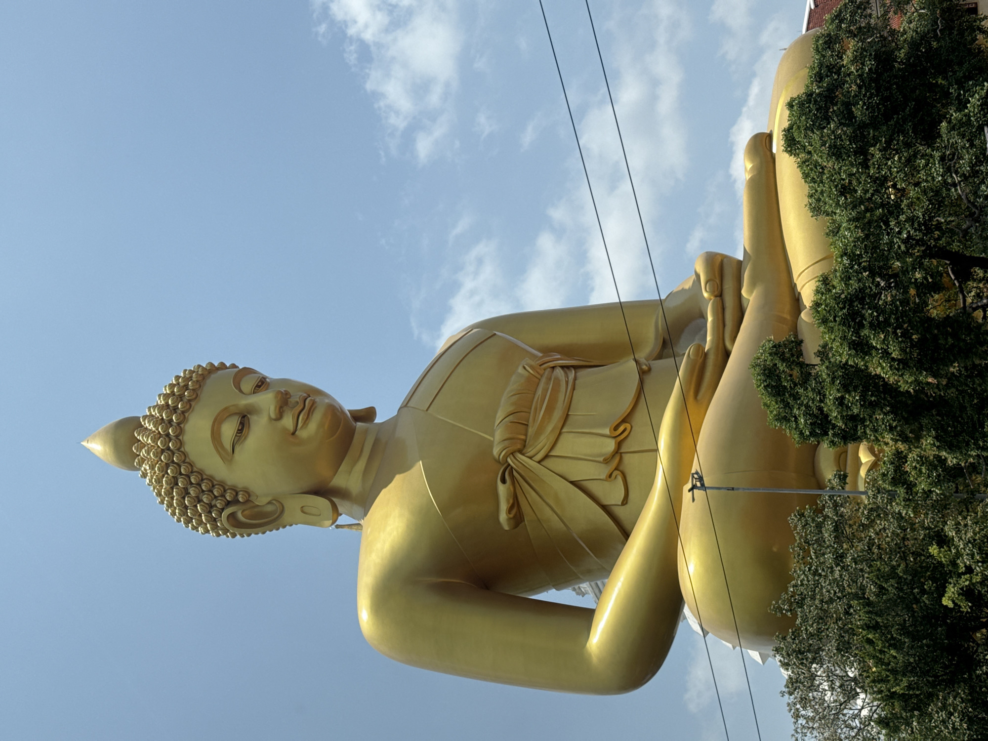 The image captures a low-angle view of a massive, golden reclining Buddha statue against a blue sky with scattered clouds. The statue is highly detailed, with intricate patterns on the head and robes. The left arm is resting on the left leg, and the right hand is in a relaxing pose. Some power lines cut across the sky above the statue. Lush green trees obscure part of the base and the upper right portion of the statue. The statue is located in Wat Khun Inthapramun, Ang Thong Province, Bangkok, Thailand.