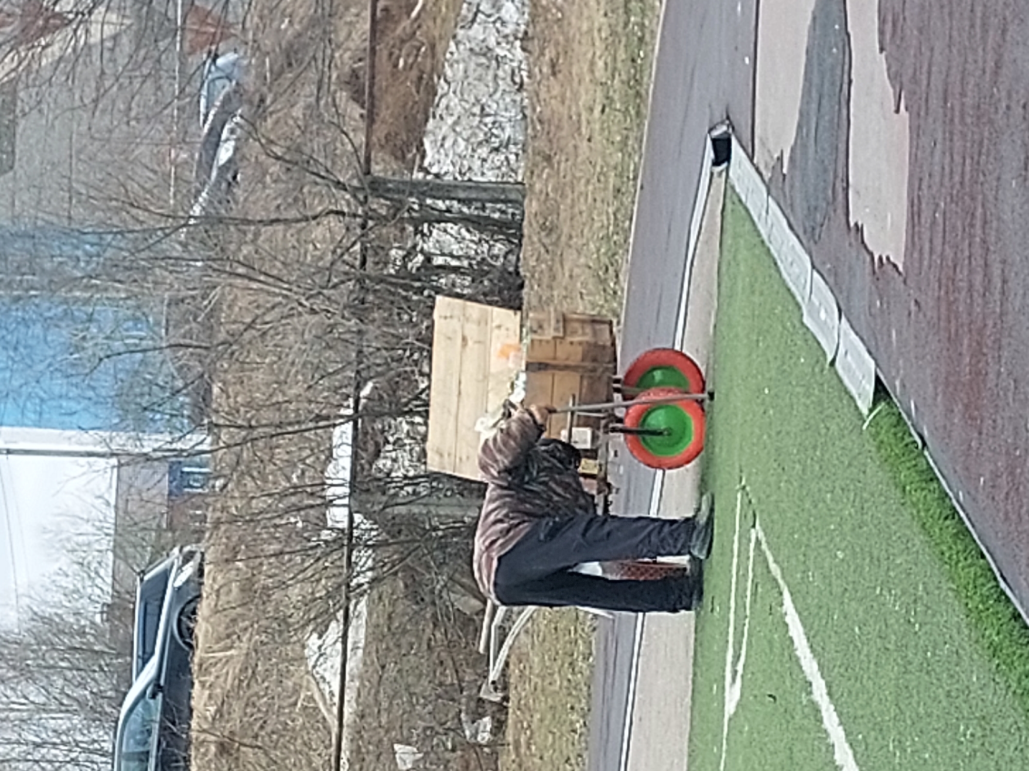A photograph taken outdoors in the city of Chaikovsky, Russia, shows a man using a roller to press white paint onto a green field. The man is wearing dark pants and a patterned jacket. The roller has orange and green components. A wooden crate and other miscellaneous objects are near the man. Trees without leaves can be seen in the background, along with a parked car. A partial view of a structure with peeling paint is also visible. The scene appears to be a field or sports area undergoing maintenance.