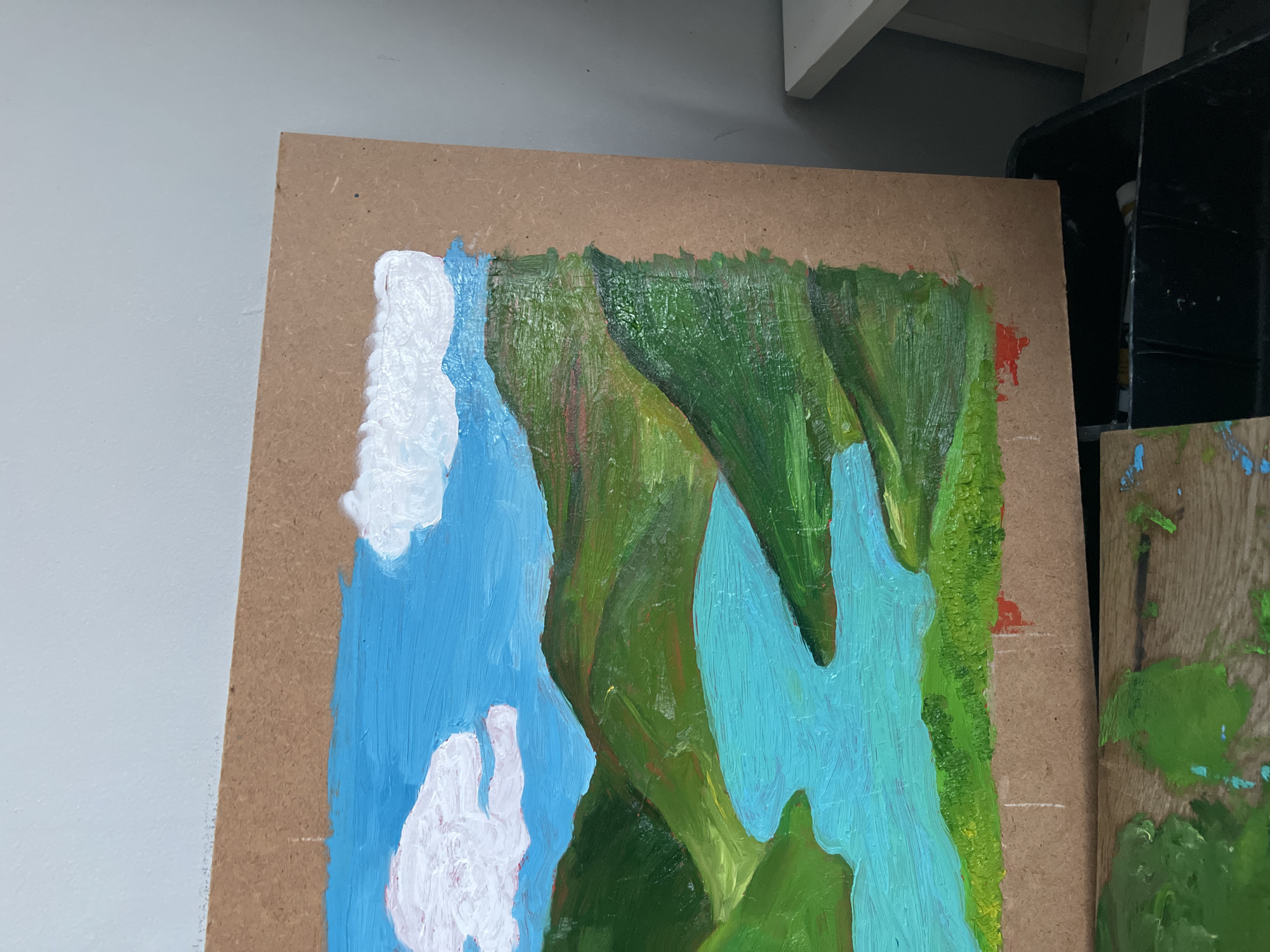 The image shows a close-up of an unfinished oil painting on a piece of wood. The painting depicts a landscape scene with blue sky and water, white clouds, and green cliffs or landmasses. The brushstrokes are visible and the paint is thick in places, particularly the white clouds. The wooden board appears unfinished, with the bare wood visible around the edges and in areas where the paint hasn't fully covered the surface. The painting is partially obscured by a black storage unit on the right and a white wall on the left. There is no indication of the city of Maidstone or its location in the United Kingdom.