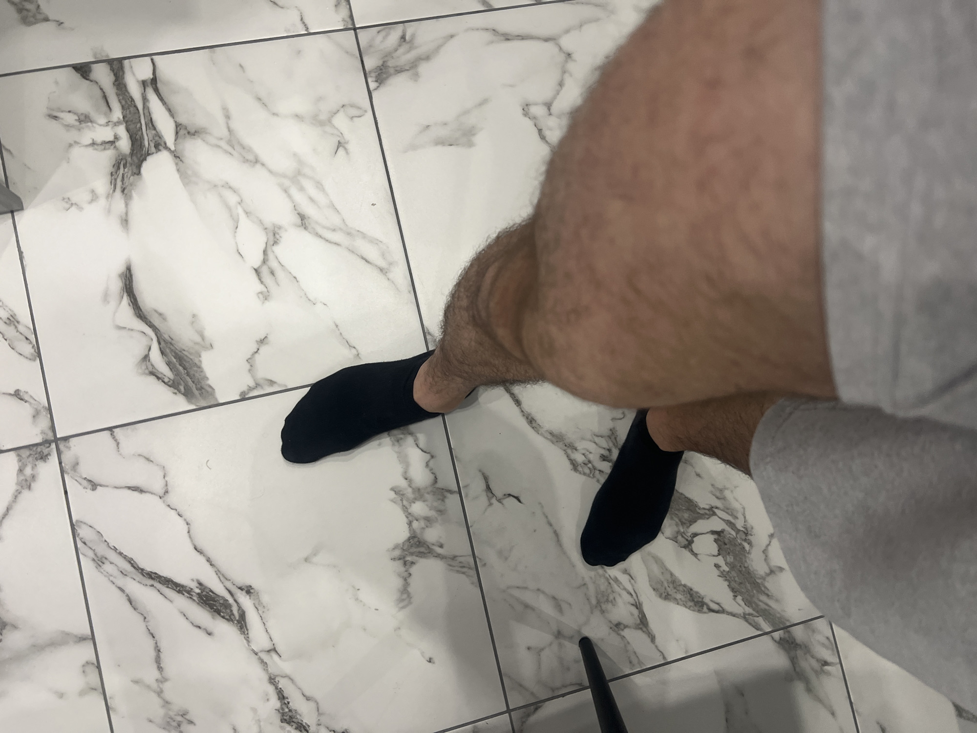 In Podgumer, Bulgaria, a person stands on a white marble-patterned tiled floor, wearing black socks and grey sweatpants. The perspective is from above, looking down at the person's legs.