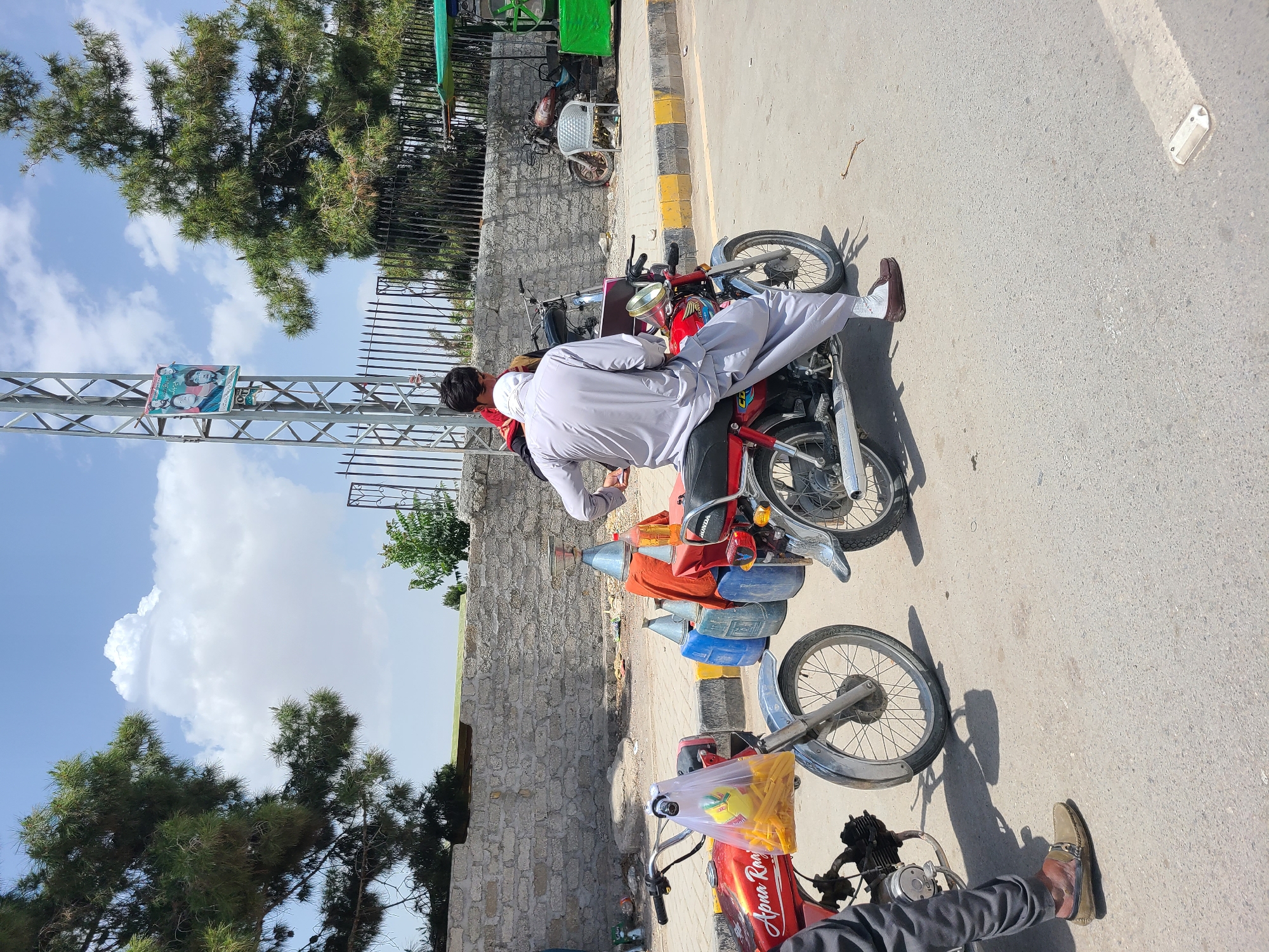 This media file captures an outdoor street scene during bright daylight in what appears to be an urban or semi-urban area, likely in Quetta City Tehsil, Pakistan.
The central subject is a man dressed in a light-colored shalwar kameez with a white head covering, seated on a red Honda motorcycle. He is positioned with his back to the camera, looking towards the right side of the frame, with his right leg extended towards the ground. The motorcycle is loaded with several plastic containers of various colors (blue, red, yellow), suggesting the transport of liquids or other goods. Another red motorcycle is partially visible in the foreground, with a clear plastic bag containing yellow items (possibly snacks or crisps) hanging from its handlebars. The brand "HONDA" is visible on the main motorcycle, and "Apna Rao" or similar text is visible on the fuel tank of the foreground motorcycle.
The scene takes place on a paved road next to a sidewalk with a distinctive yellow and black striped curb. A sturdy stone wall runs parallel to the road, topped by a black metal fence. Beyond the wall and fence, tall green trees with needle-like foliage are prominent against a clear blue sky with scattered white clouds, indicating sunny weather. Strong shadows cast by the subjects and trees suggest it is mid-day or early afternoon. A metal light pole stands in the mid-ground, bearing a political poster that features faces of a woman and several men, along with Urdu script. In the background, a small green structure or vehicle (possibly a rickshaw or cart) is visible near the wall. The overall impression is a bustling roadside environment.