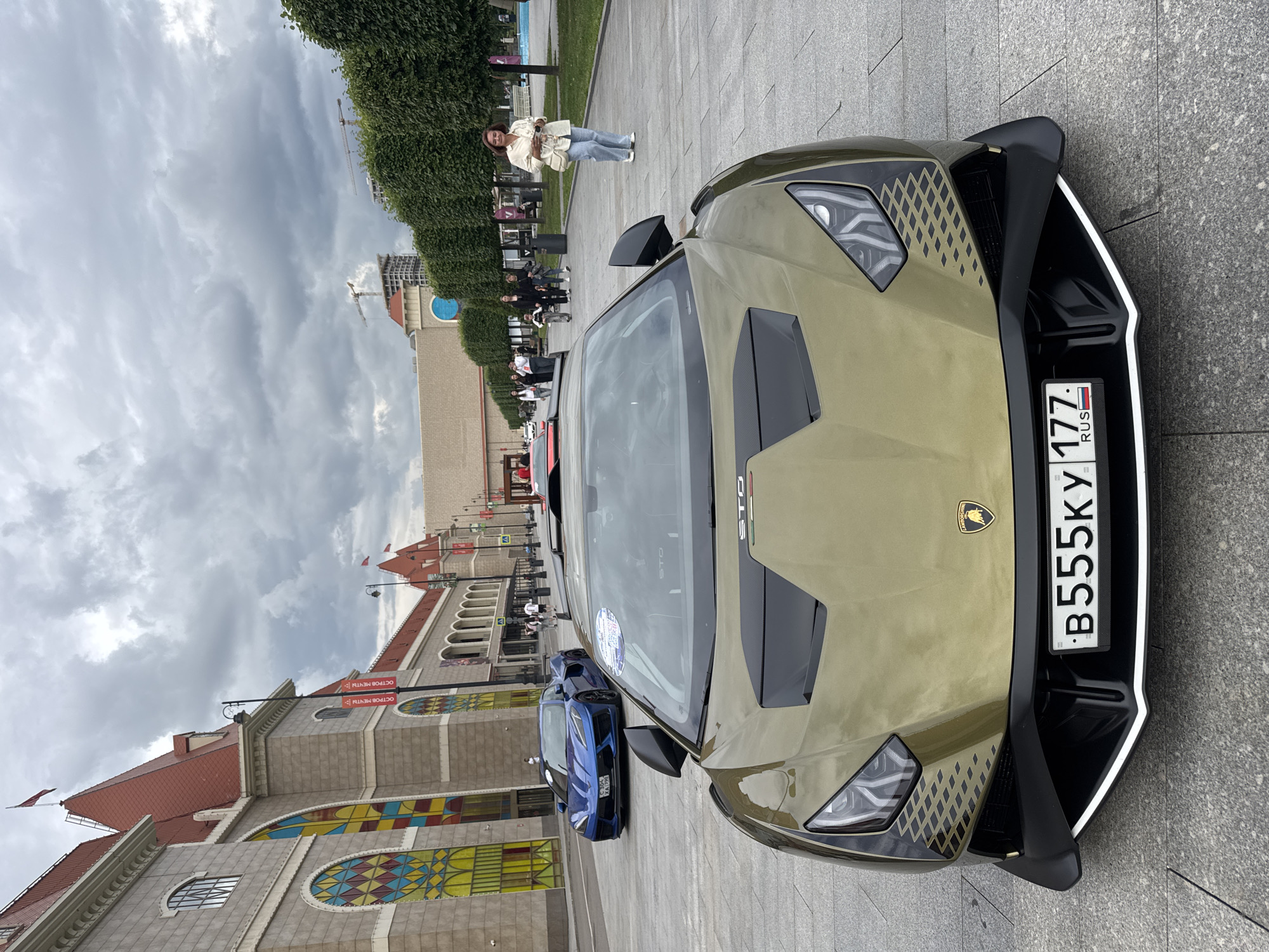 The image shows a gold Lamborghini Huracán STO parked on a paved plaza in front of a building with a red roof and colorful stained-glass windows. A blue Chevrolet Corvette is parked further down the plaza. Several people are walking in the background. The building appears to be part of an entertainment complex, likely Ostrov Mechty (Dream Island) in Moscow, Russia, given the presence of multiple Russian license plates. The sky is overcast, and trees line the right side of the plaza.