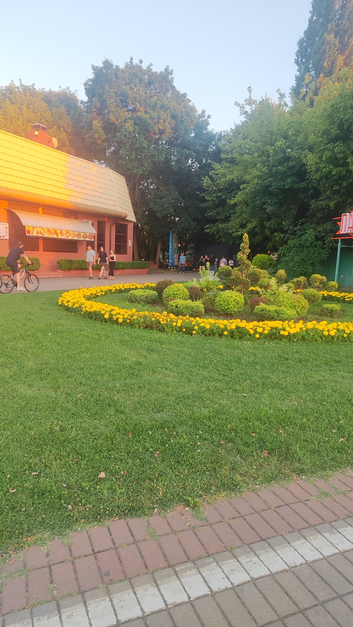The image depicts an outdoor scene with a building on the left and a garden in the center and right. The building has a red facade and a yellow tiled roof. Several people are seen walking near the building and a man is riding a bicycle in the foreground.
In the center of the image is a landscaped garden with a circular flower bed filled with yellow flowers. Behind the flowers, there are trimmed bushes and a tall, conical topiary. Further to the right, a green structure with a red sign that says "ПАРК" (PARK in Russian) is visible.
The area is covered with green grass and a paved walkway made of red and grey bricks in the foreground. Tall trees with lush green foliage surround the scene, and the sky is clear and light blue. The lighting suggests it is either early morning or late afternoon, with a warm glow on the building.
This scene is located in the park "Sunny Island" in Krasnodar, Russia. The building on the left is likely a cafe or an ice cream parlor within the park, and the "ПАРК" sign indicates the entrance or a zone within the amusement park area.