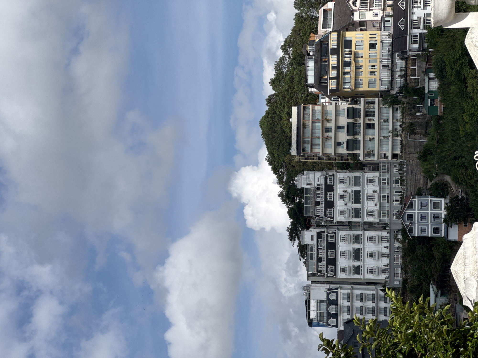 The image depicts a section of Da Lat, Vietnam, characterized by a dense collection of European-style buildings situated on a hillside. The buildings, varying in architectural design and color, are multi-storied with prominent features like pitched roofs, balconies, and numerous windows. Some structures display white facades with black trim, while others are painted yellow or feature darker grey exteriors. The buildings are set against a backdrop of lush green trees and a sky filled with scattered white clouds. The composition suggests a high vantage point, looking down upon the urban landscape and the surrounding natural elements. The overall impression is one of a densely populated, hilly area with a blend of residential and commercial architecture.
