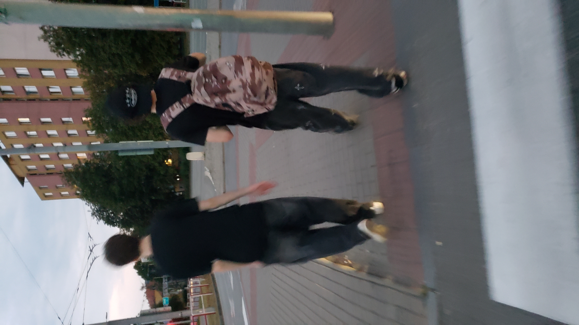 Two individuals are walking on a sidewalk in front of a multi-story building. The person on the left is wearing a black t-shirt and dark pants, with light-colored shoes. The person on the right is wearing a black t-shirt, dark pants with a camouflage backpack, and dark shoes. The building is light pink and yellow with numerous windows. Trees are visible behind the building. The sidewalk is paved with rectangular stones. A crosswalk is visible in the foreground. The scene appears to be in a city setting, possibly Prešov, Slovakia, given the urban architecture and infrastructure. The lighting suggests it is either early morning or late evening.