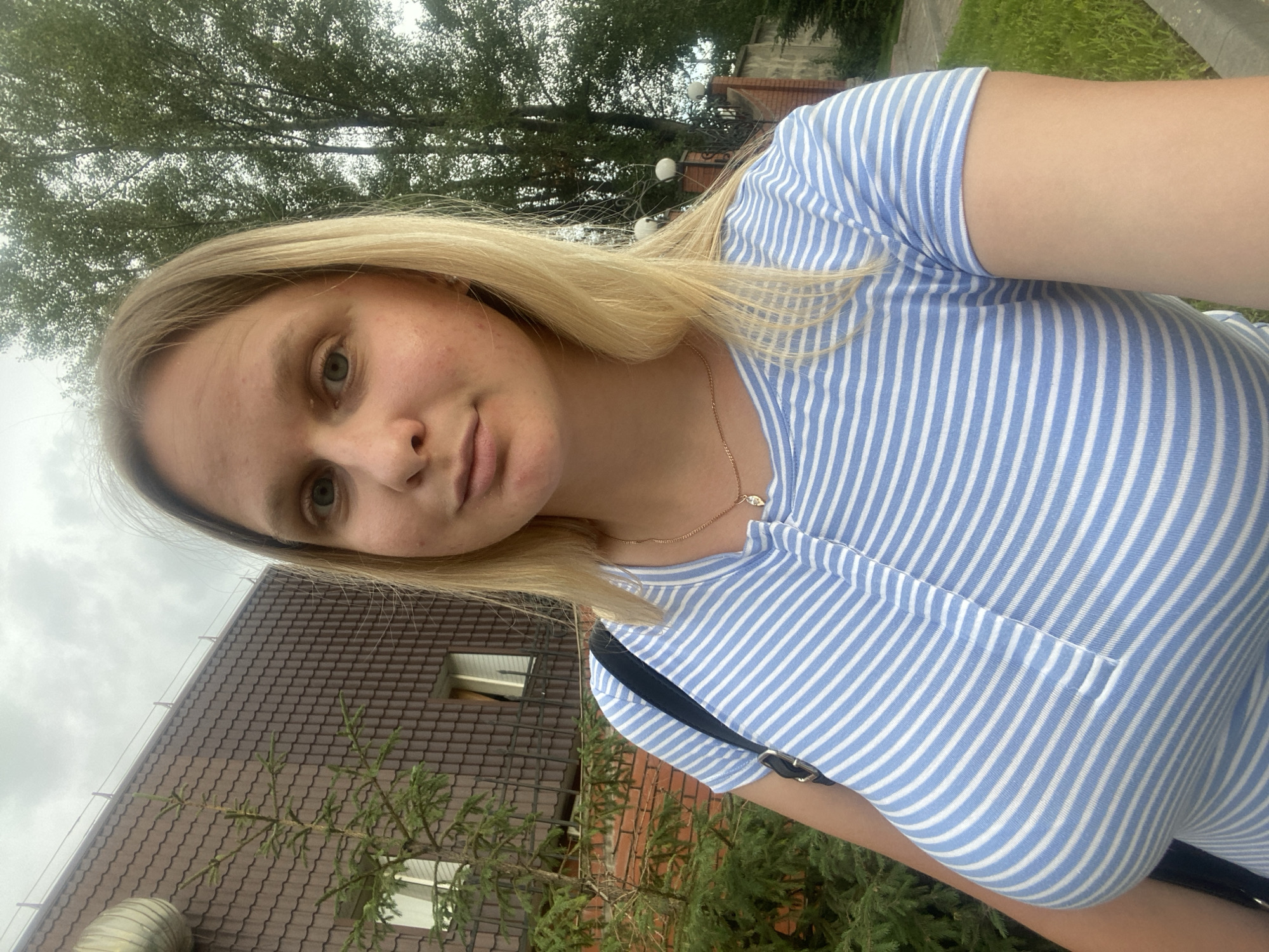 In the city of Chaikovsky, Russia, a young woman with blonde hair and fair skin takes a selfie. She is wearing a blue and white striped t-shirt and a delicate gold necklace. The background shows a brown-tiled building, a green tree, and a cloudy sky. The woman's face is slightly turned towards the camera, and she has a neutral expression. The image captures a moment of everyday life in an urban setting.