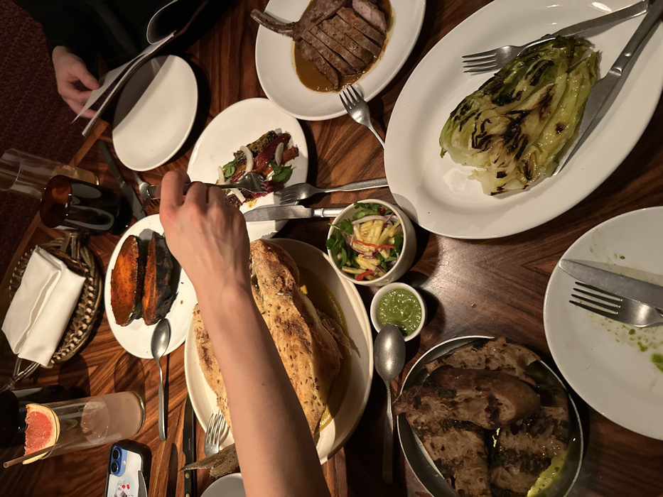 An overhead shot captures a lively dinner scene at a restaurant. Several hands are visible around a dark wooden table, laden with various dishes, suggesting a shared meal among friends or family. One person's arm and hand are prominent in the foreground, holding a fork over a large oval dish containing what appears to be a baked fish or large herbed bread loaf, partly submerged in a yellow sauce. To the left, another person's hands are seen holding an open menu or document.
The table is filled with an array of food. Main dishes include a plate with neatly sliced meat, possibly a pork chop, drizzled with a rich brown sauce, and another plate featuring a grilled head of lettuce, exhibiting char marks. Side dishes comprise a serving of roasted sweet potato halves, a small bowl of fresh green salad with mango and red onion, and another small bowl with a vibrant green dip or sauce. A basket holds slices of grilled bread, and another plate holds a darker salad with mixed ingredients.
Beverages include a dark glass carafe or bottle, and a clear glass cocktail adorned with a slice of grapefruit and a straw. A white-cased mobile phone, possibly an iPhone, with a small decorative sticker is placed near the cocktail. The tableware consists of white plates, forks, knives, and spoons. The lighting is dim and warm, indicating an evening setting. The overall atmosphere suggests a social and enjoyable dining experience. No legible text is visible on the menu or phone.