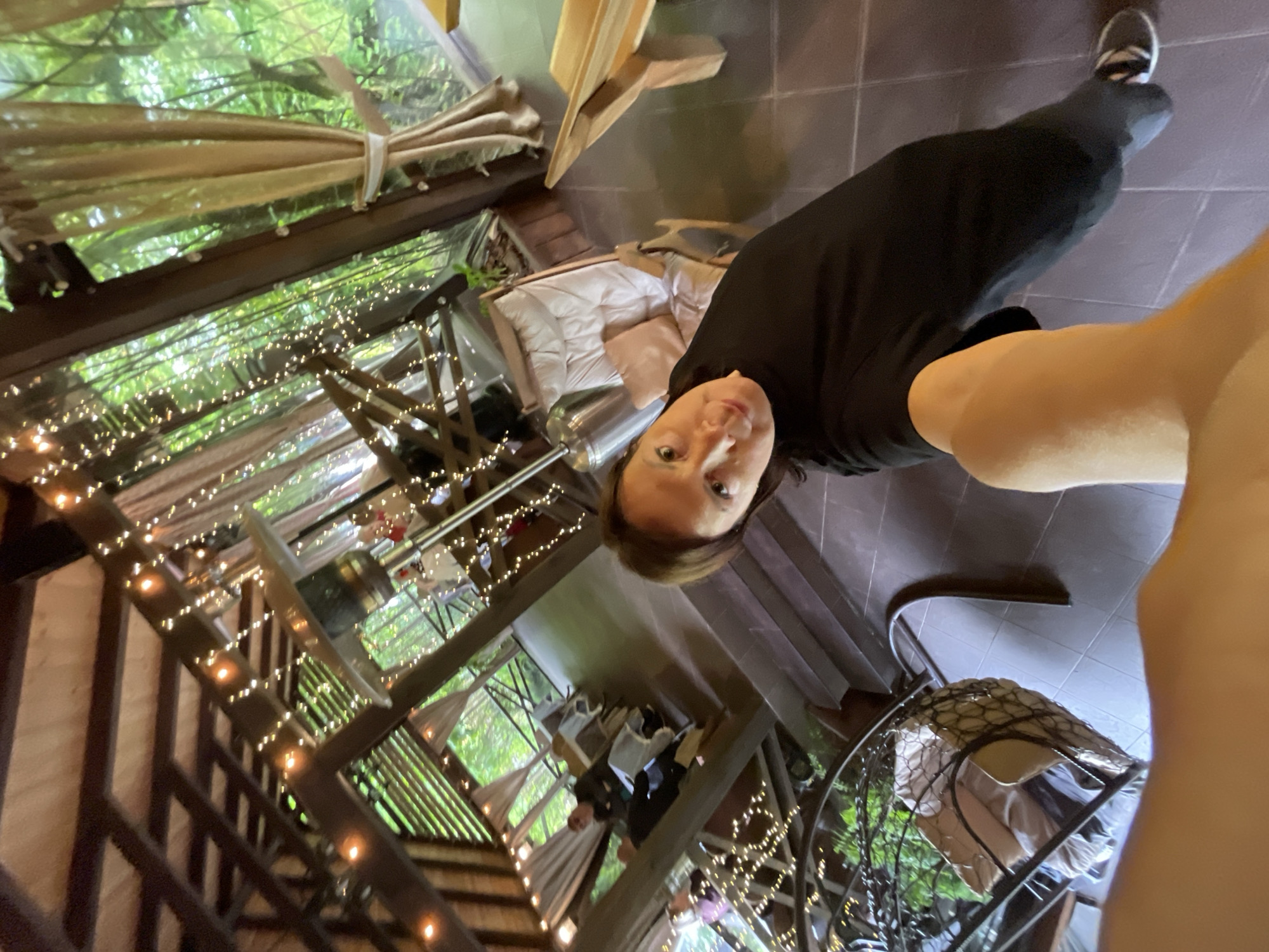A high-angle perspective photograph shows a woman in Solokh-Aul, Russia, in what appears to be an outdoor or partially enclosed space. The woman is wearing a black top and bottoms with sneakers. The location features string lights and wooden beams, suggesting a deck or similar outdoor structure. There is furniture, including chairs and what seems to be outdoor seating with cushions. The view includes greenery, which suggests an outdoor setting integrated with nature.