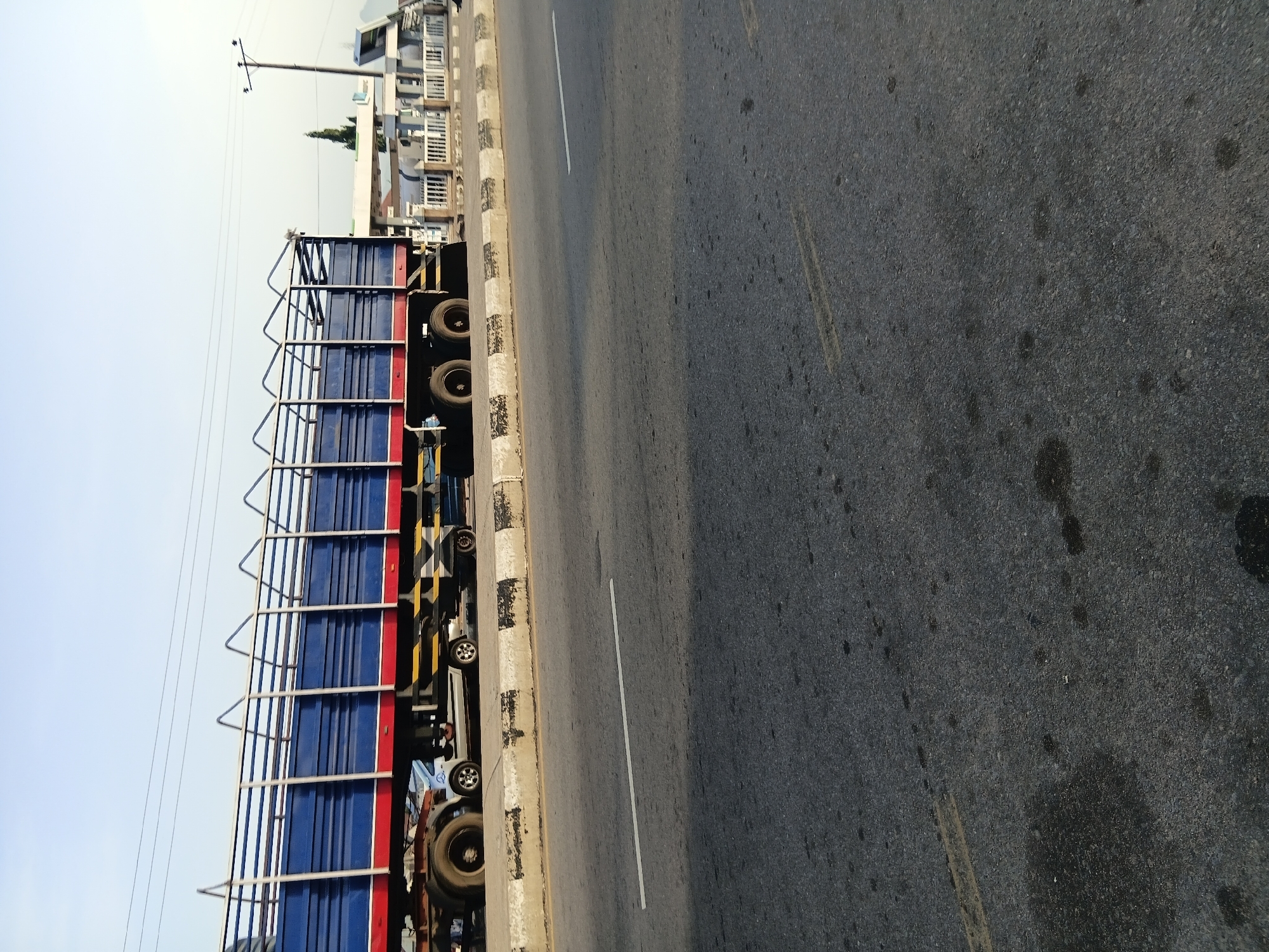 A wide-angle, eye-level shot captures a large truck with a blue and red cargo bed and a metal cage enclosure parked on the side of a road. The truck is positioned horizontally across the frame, with its wheels visible. The road appears to be asphalt, with visible tire marks and dark stains. A concrete curb, painted with alternating black and white stripes, separates the road from a sandy area. In the background, a white building with a fence and a utility pole with wires are visible. The sky is clear and pale blue, suggesting it is daytime. The scene is likely in Jalingo, Nigeria, given the context provided.